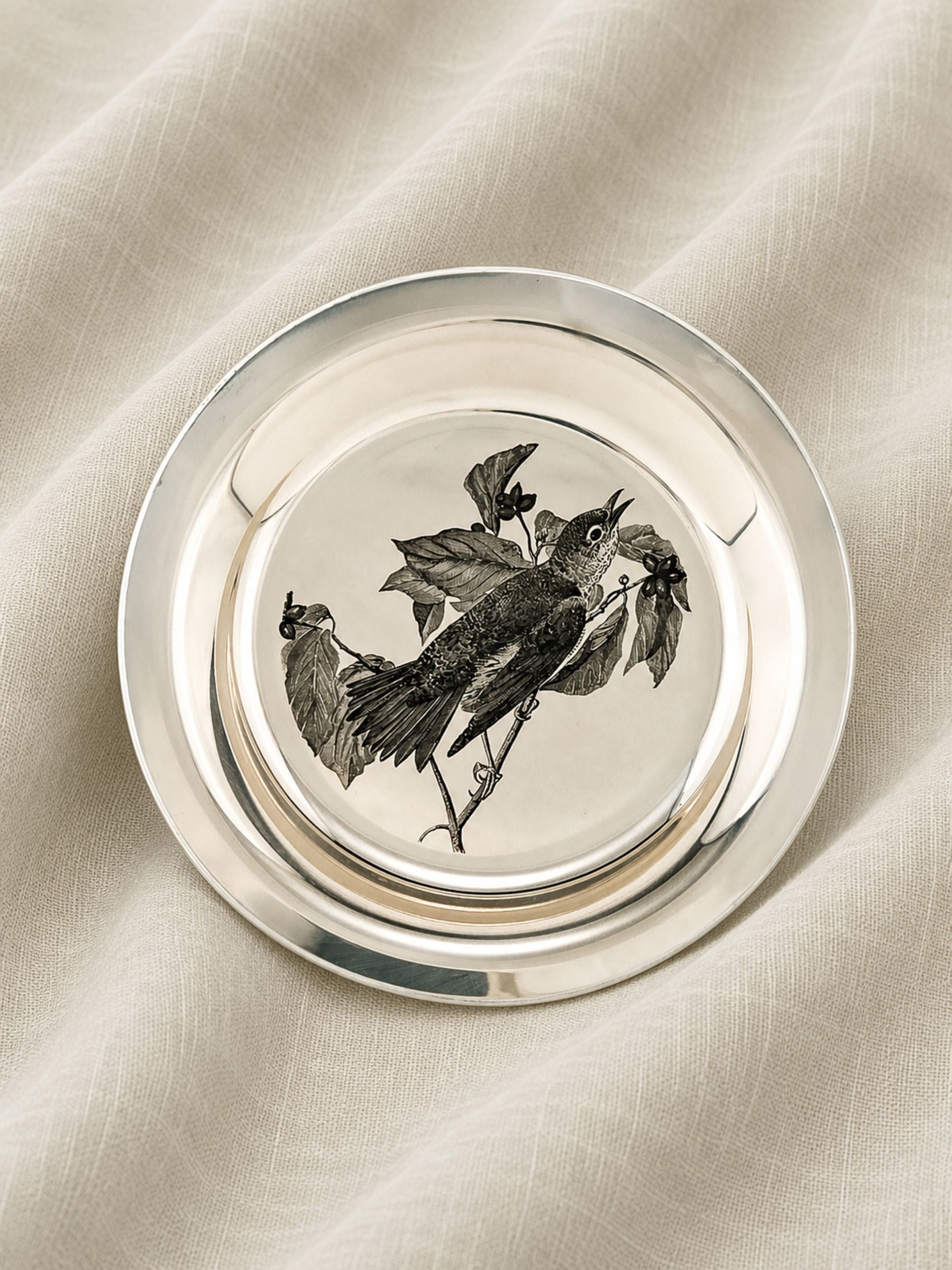 A round Sterling silver plate with a raised rim, featuring a black-and-white illustration of a wood thrush birds on branches in the center, photographed on draped cream fabric.