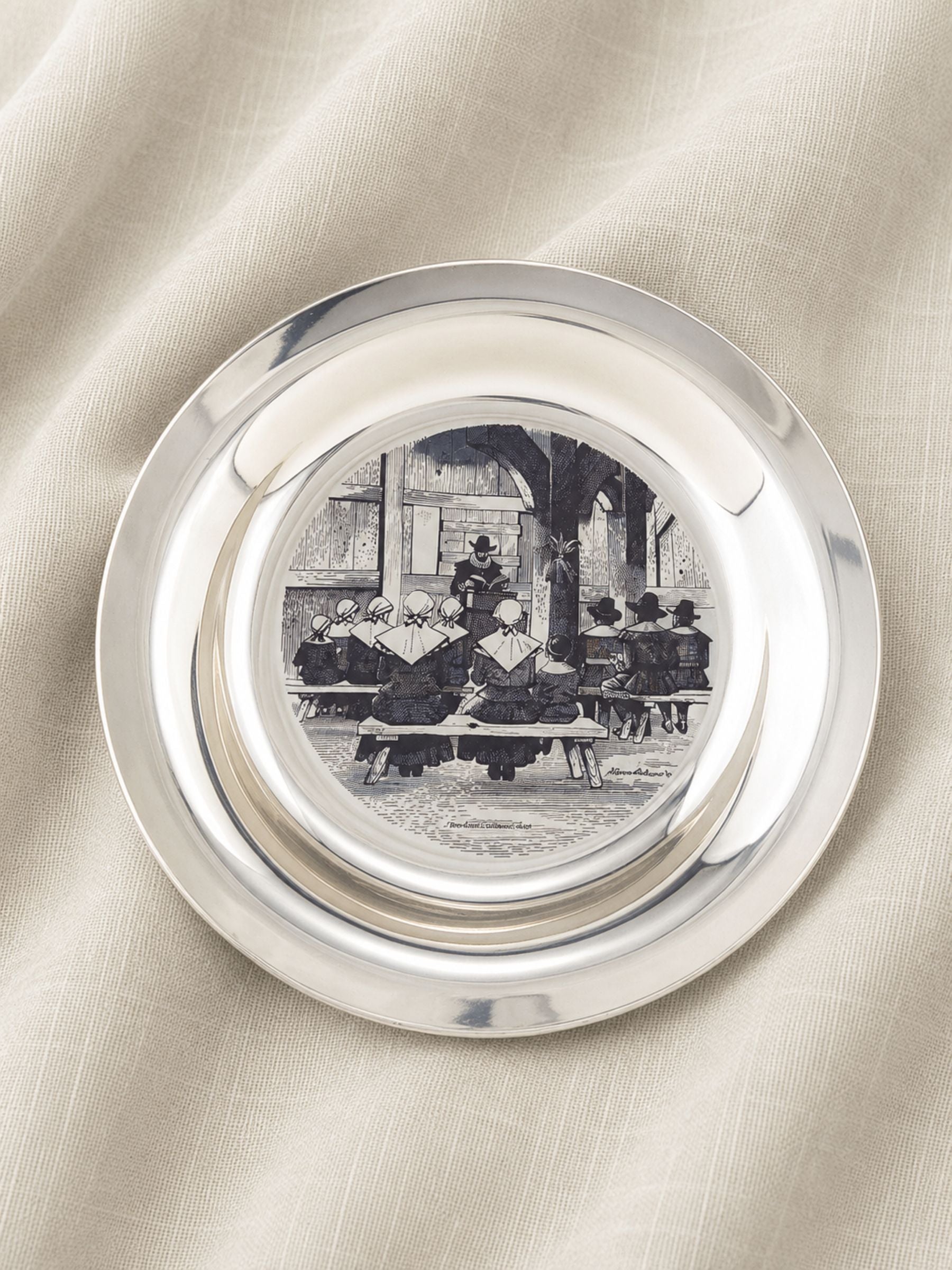 A round silver-toned decorative plate by Stevan Dohanes with a central black-and-white printed scene of people seated at a table, photographed on a draped cream fabric background.
