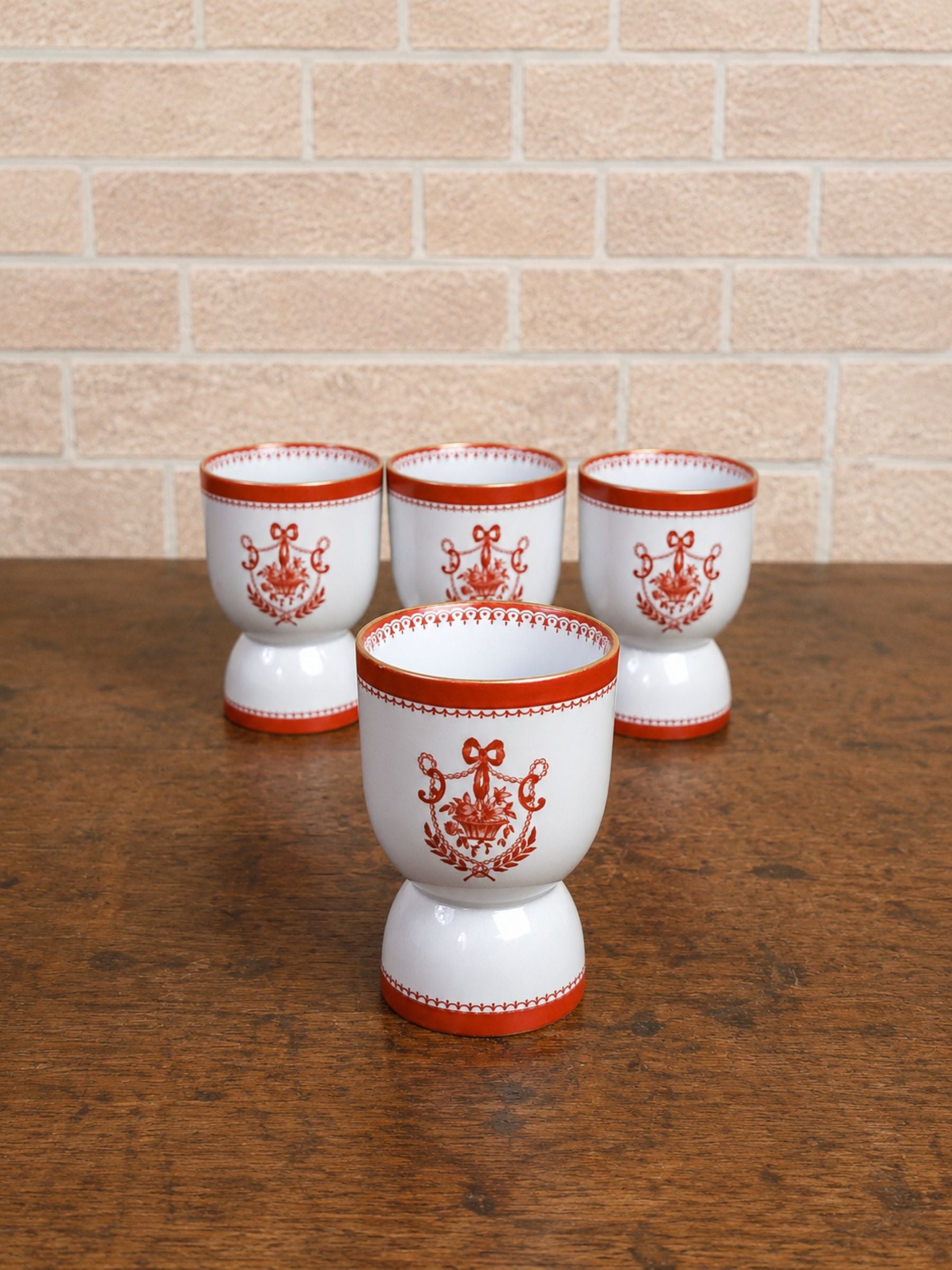 Four gray ceramic small goblets with red decorative patterns and borders, arranged on a wooden tabletop against a light brick wall background.