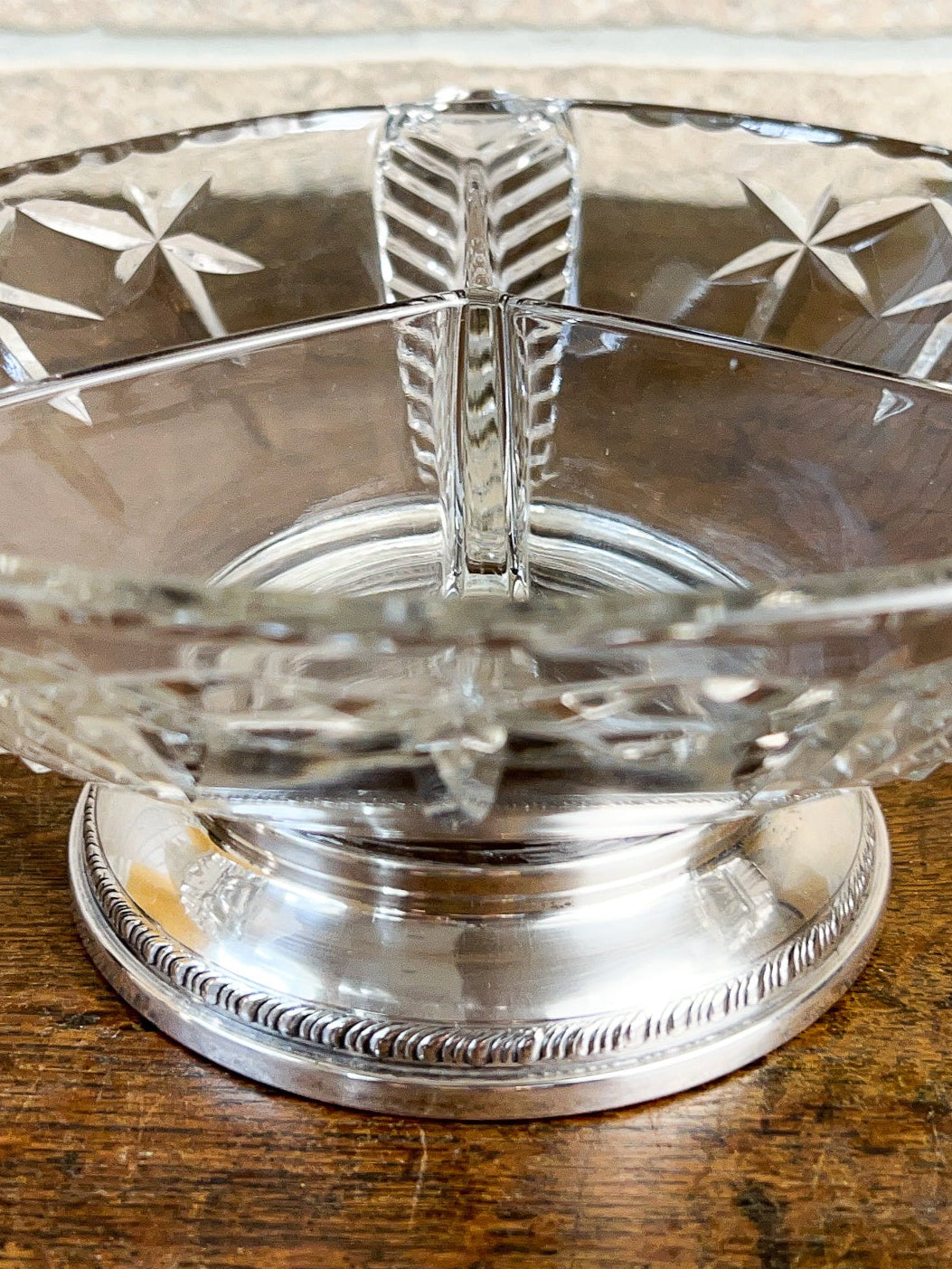 A clear cut-glass pedestal bowl with a sterling silver base set on a wooden surface against a light brick wall.