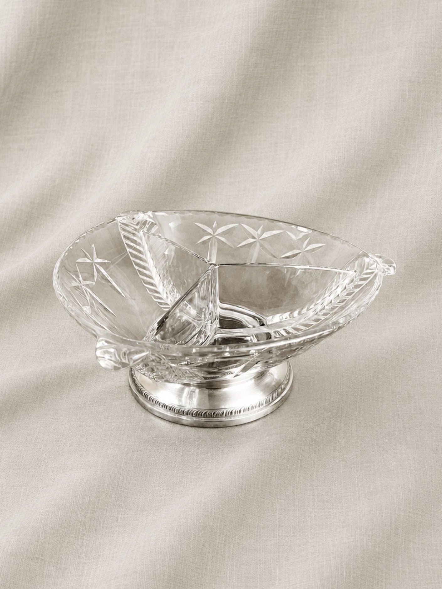 A clear cut-glass oil lamp with a silver-toned metal base, set against a draped beige fabric background.