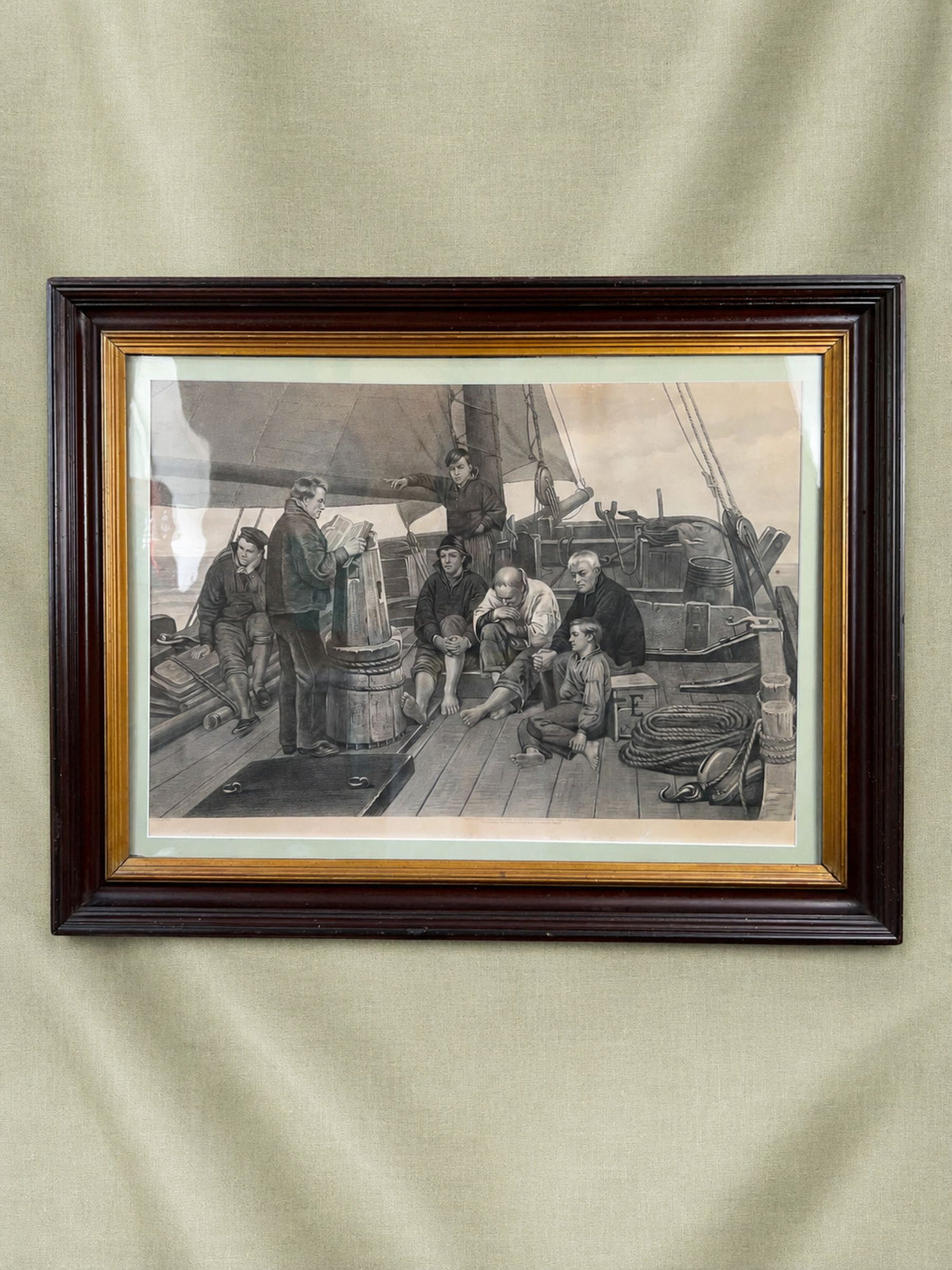 A framed antique sepia-toned print depicting a group of men on a ship's deck, enclosed in a dark brown wooden frame with a gold inner trim and a cream mat, hung against a textured beige fabric backdrop.