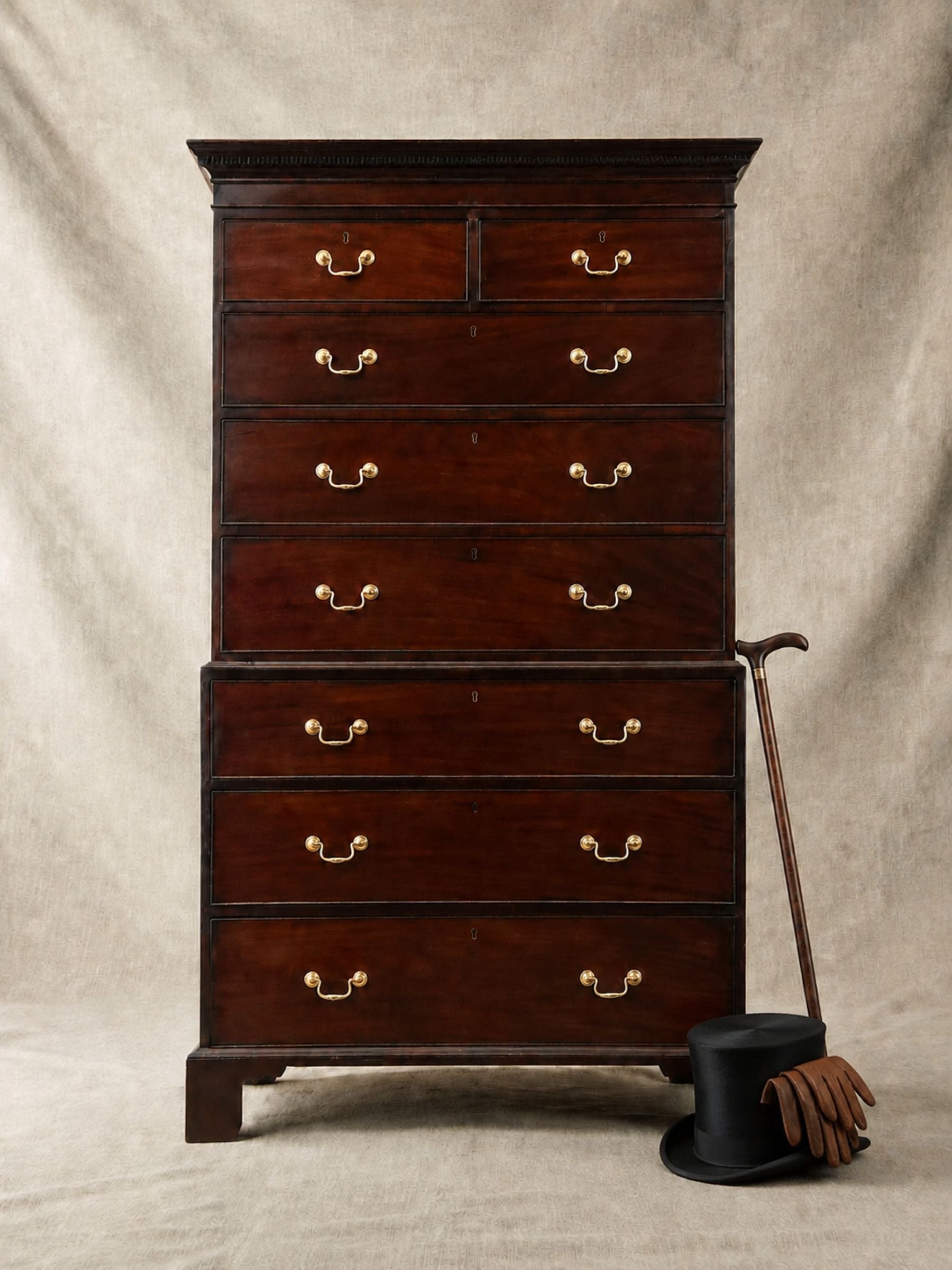 A tall dark wood chest of drawers with brass handles, photographed against a neutral draped backdrop. A cane, black top hat, and brown gloves are placed beside it on the floor.