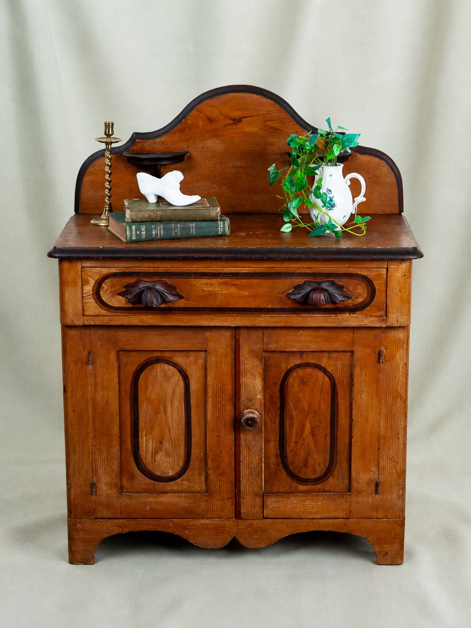 An antique wooden chest of drawers or side cabinet with a curved back panel, featuring a single drawer with decorative dark handles and two lower doors with oval panels. The cabinet is styled with books, a small white sculpture, a brass candlestick, and a white pitcher with green leaves on top, set against a draped beige fabric backdrop.