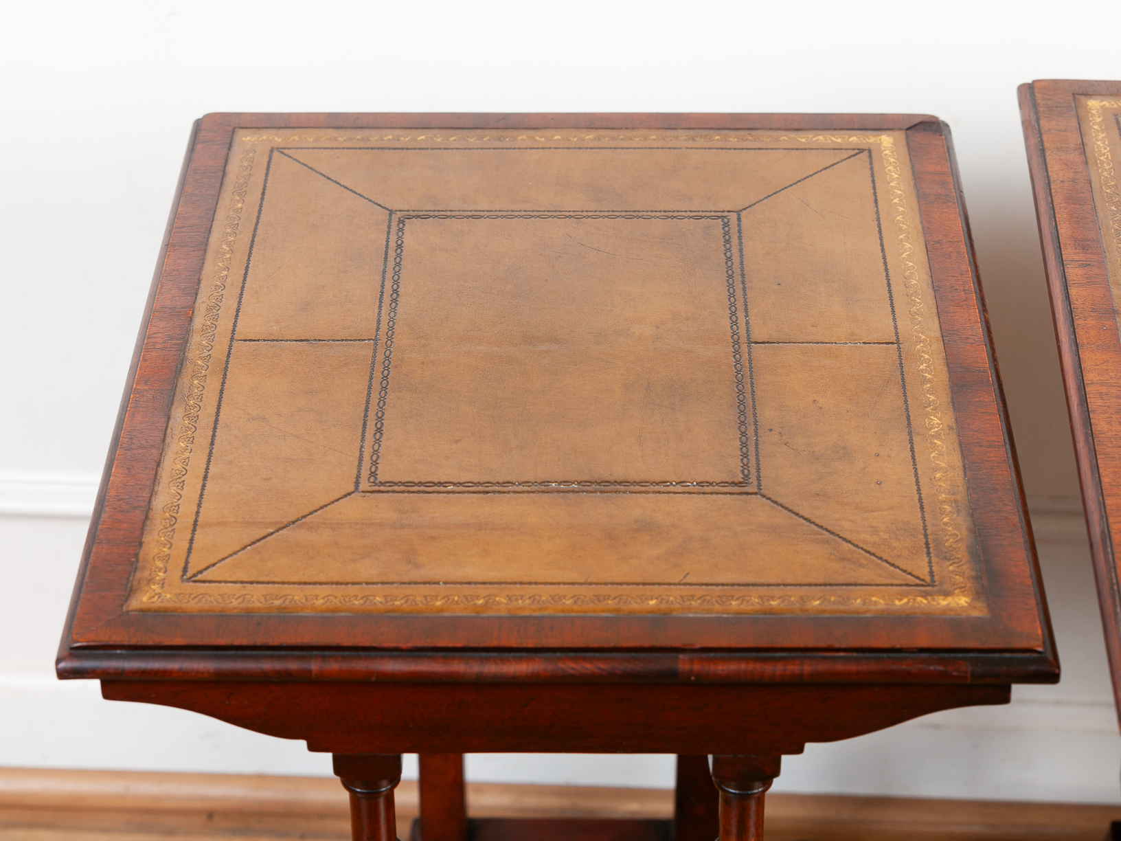 A close-up of a wooden tabletop with a warm olive leather inlay bordered by dark wood and gold-toned decorative lines, shown against a white wall.