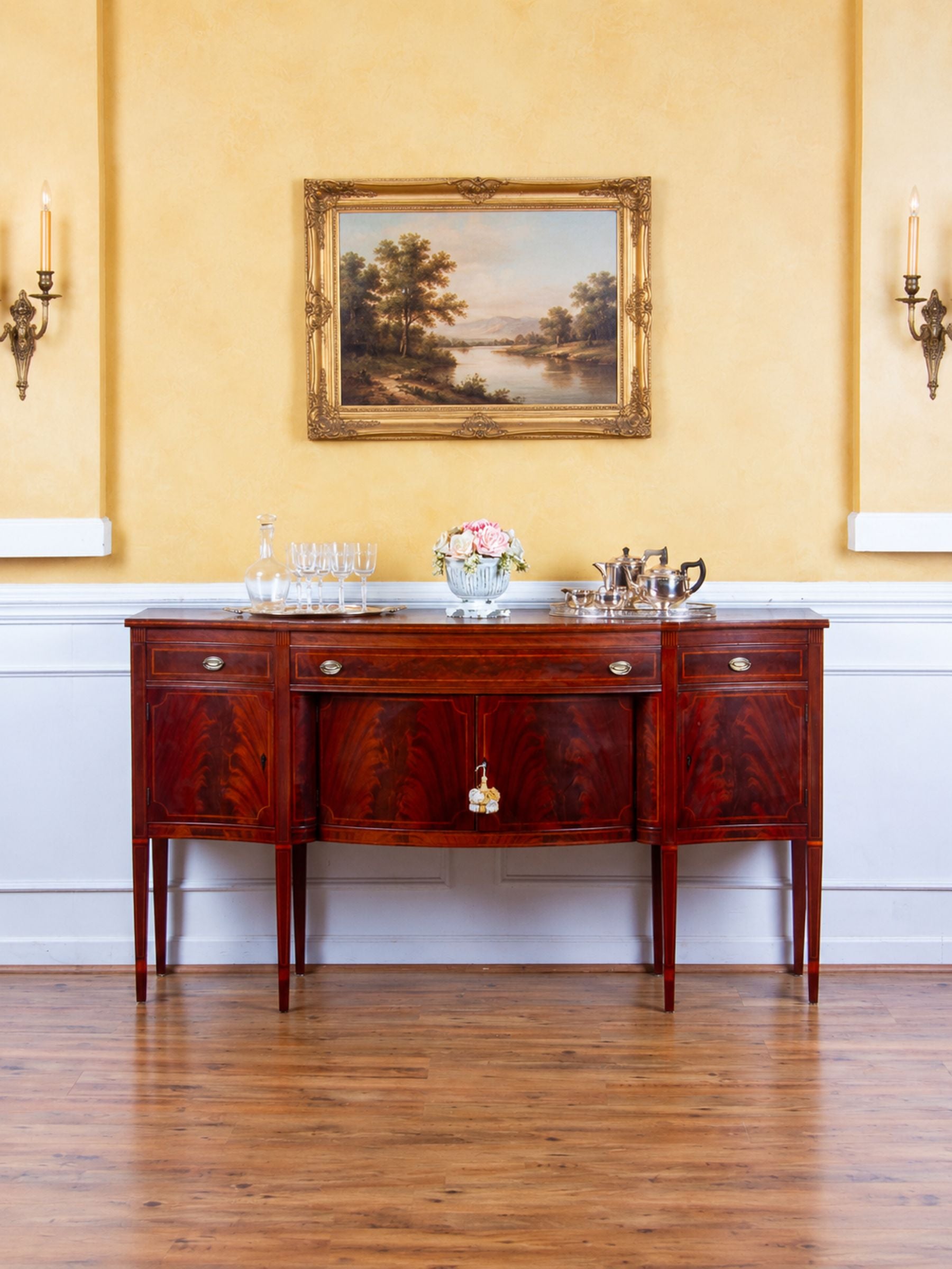 A classic mahogany sideboard console table with a polished dark reddish-brown mahogany wood finish, set against a yellow wall with a framed landscape painting and wall sconces.
