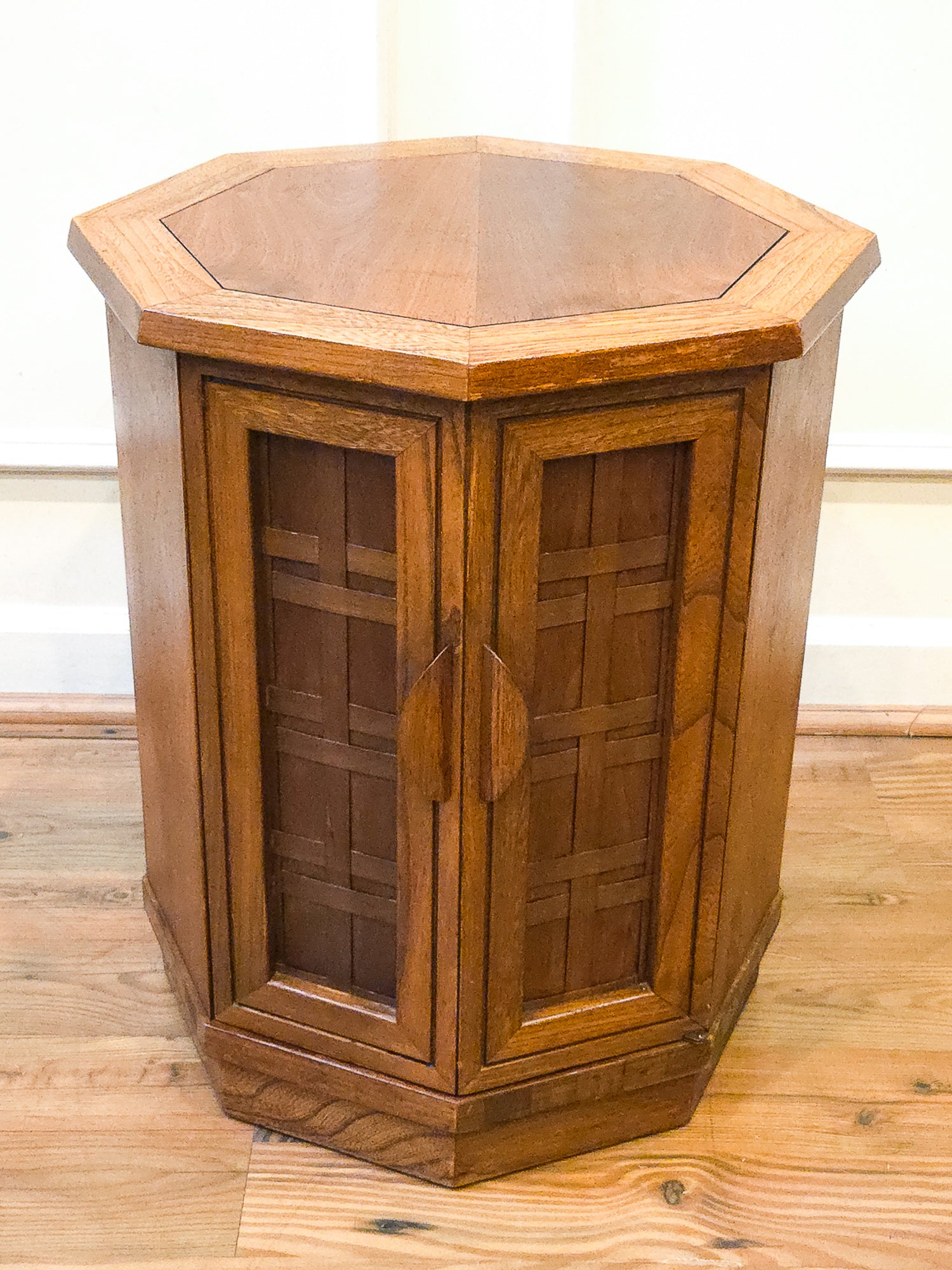 A hexagonal wooden cabinet or side table with two paneled doors and a hexagonal top, shown on a wood floor against a light wall.