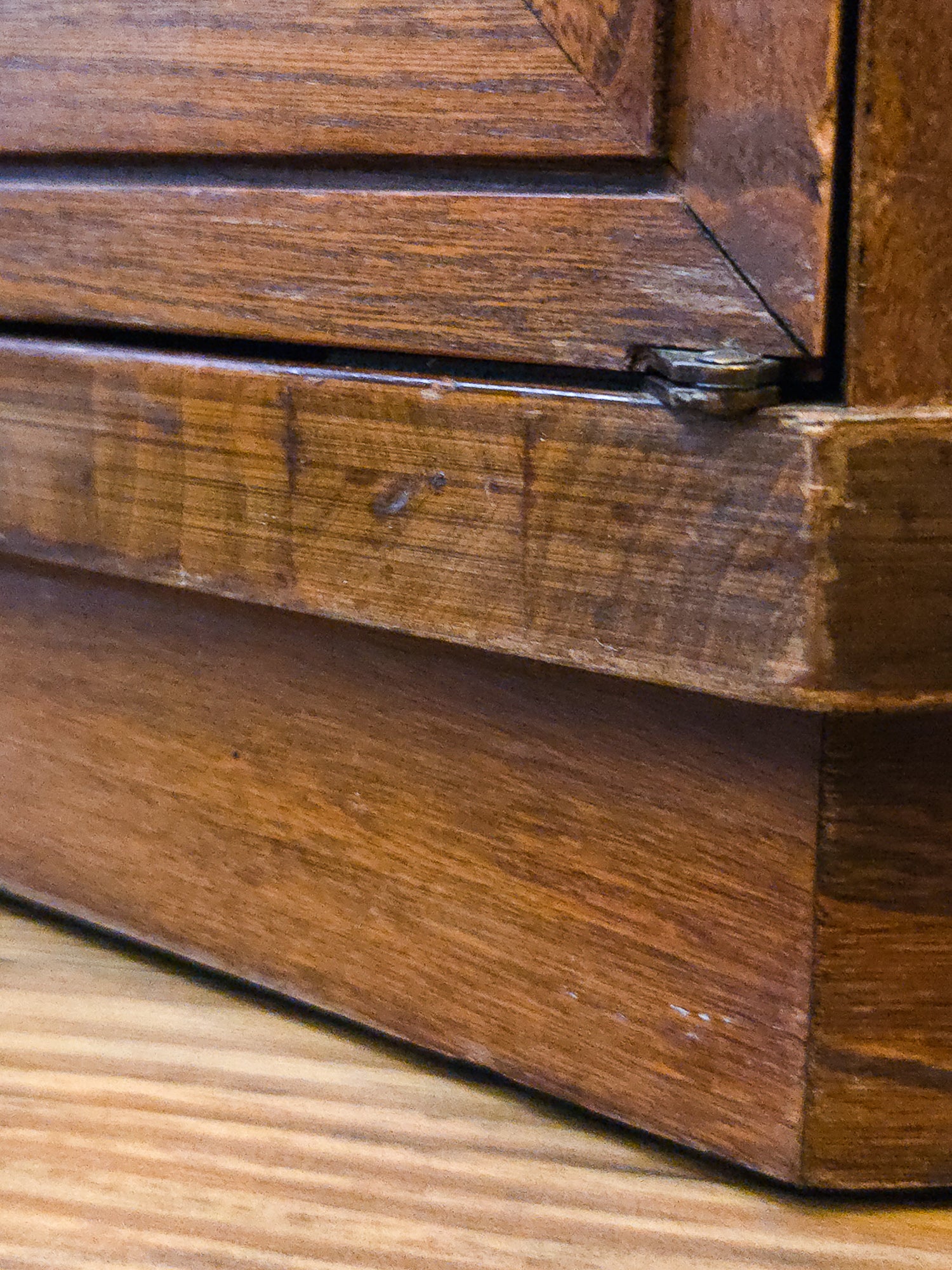 Close-up of a dark brown wooden furniture piece, showing the wood grain, corner joint, and a small metal hinge or latch, resting on a light wood floor.
