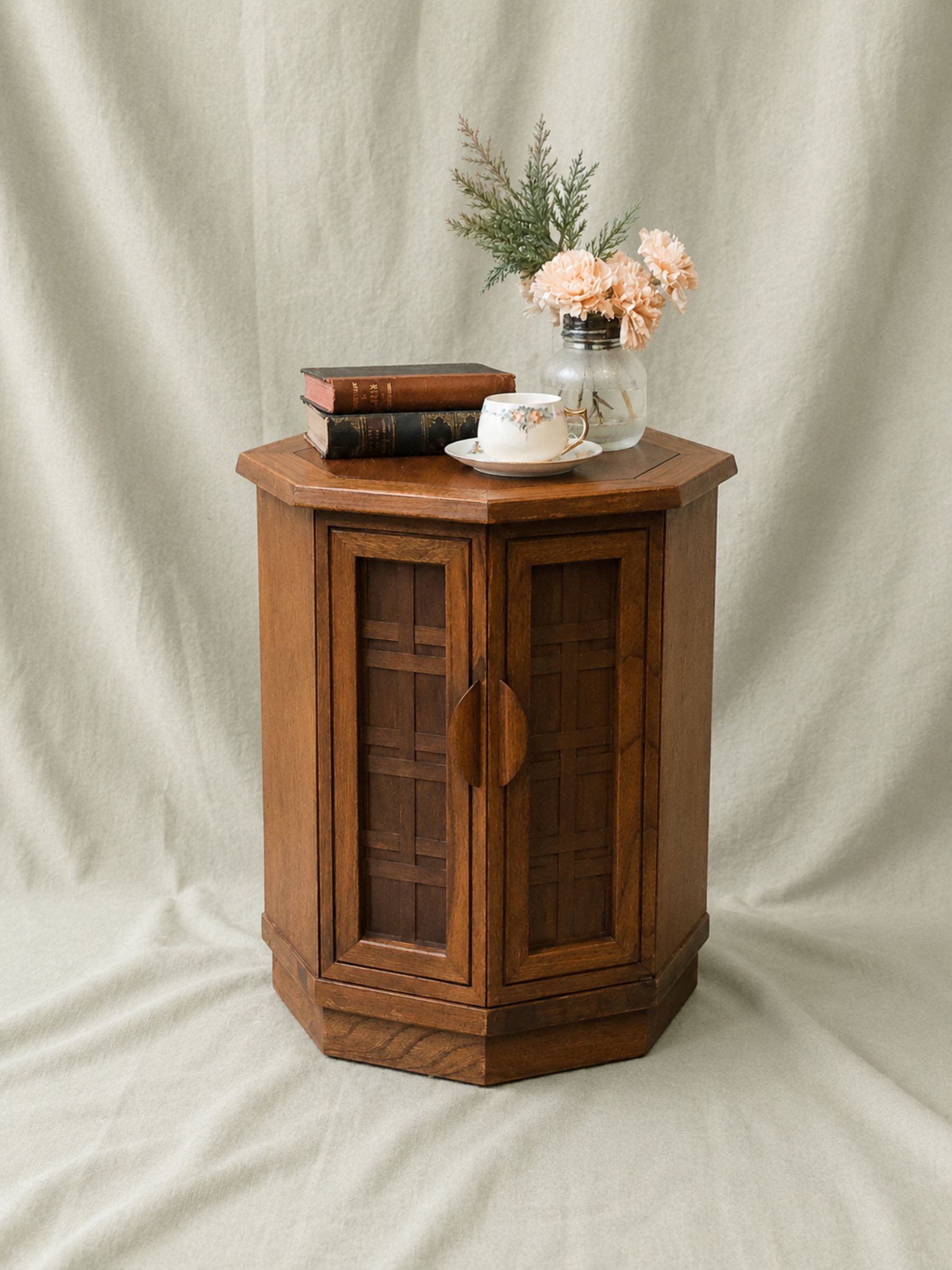 A hexagonal wooden side cabinet with two glass-paneled doors, styled on a draped beige fabric backdrop with books, a teacup, and a glass vase of flowers.
