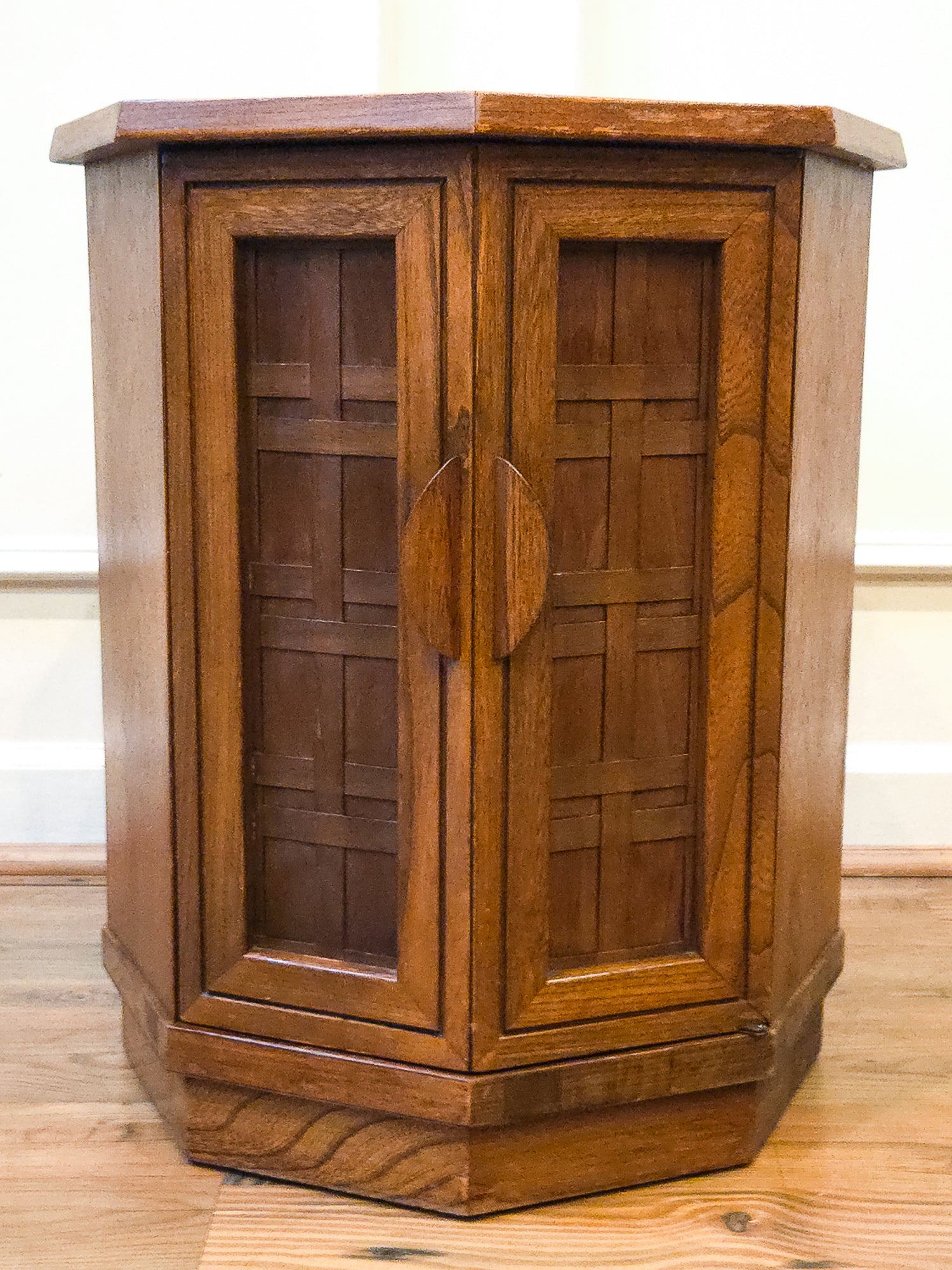 A vintage octagonal wooden cabinet with two doors featuring geometric paneled designs and oval handles, set on a wooden floor against a white wall.