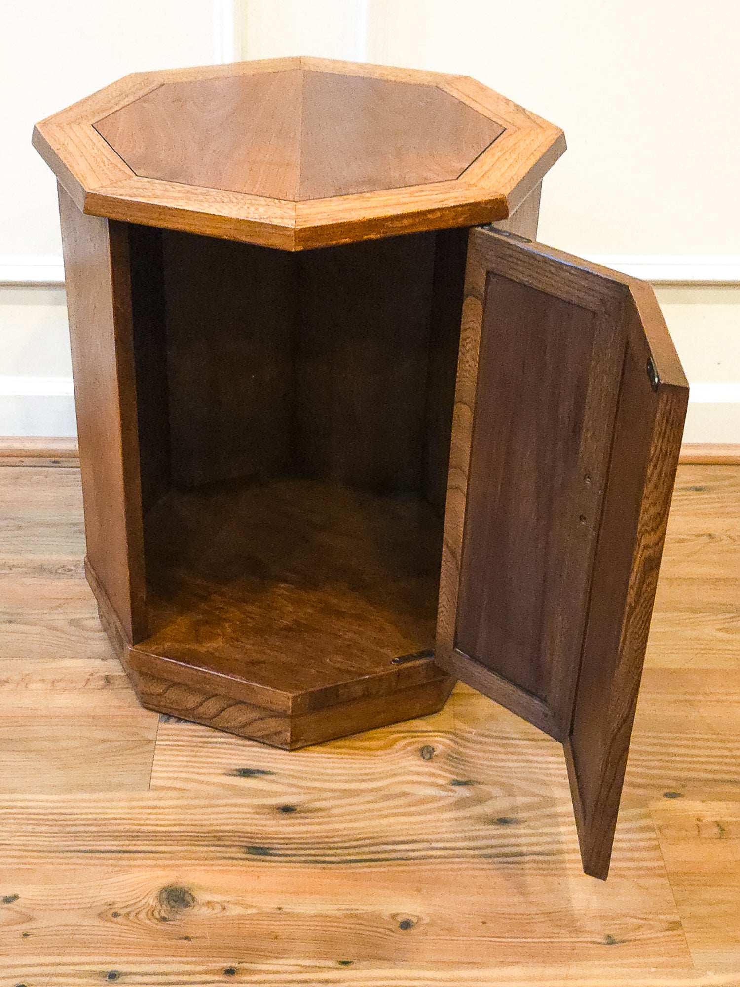A hexagonal wooden storage cabinet with an open door, showing a dark wood interior, set on a light wood plank floor against a white wall.