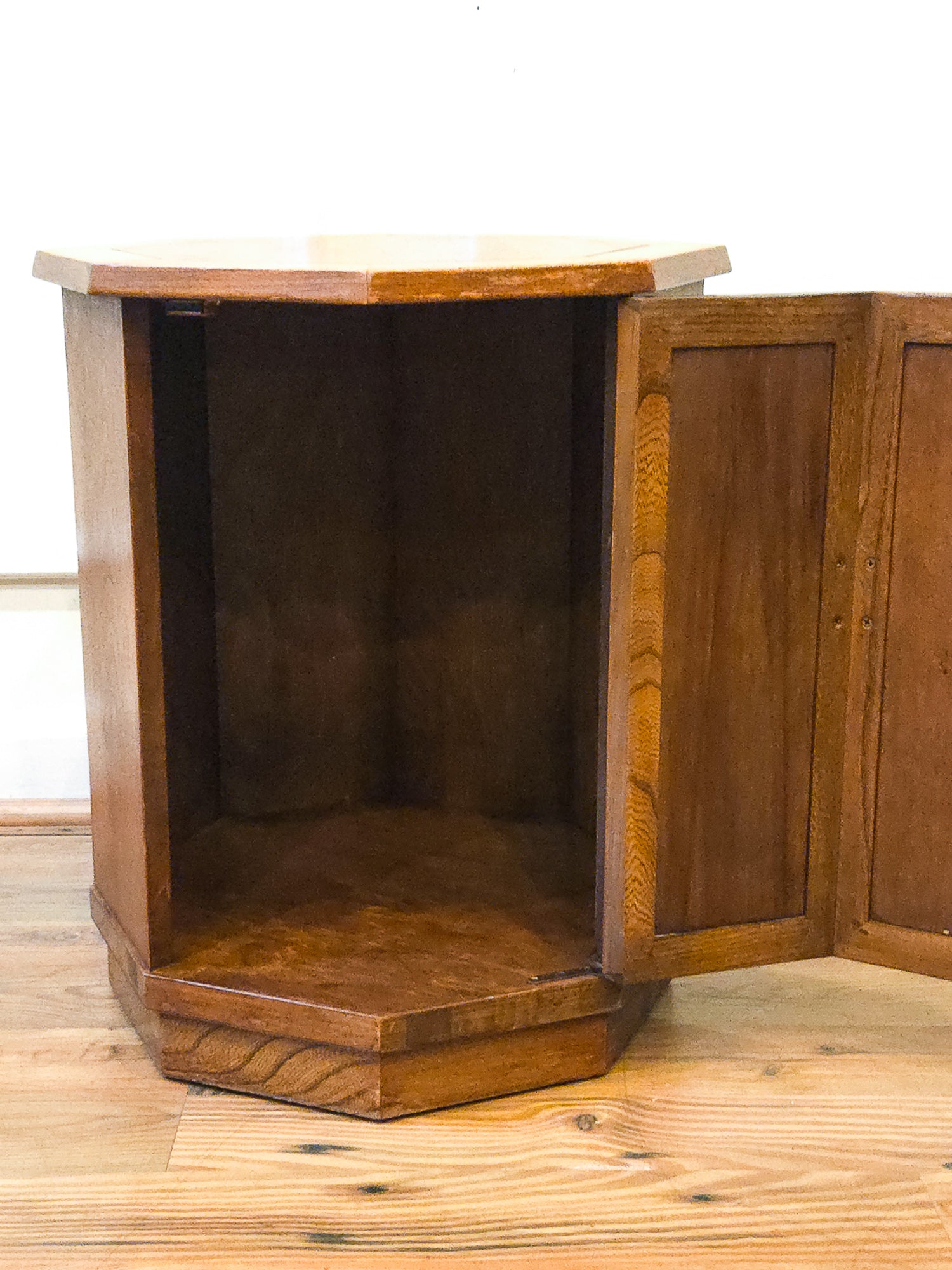 A hexagonal wooden side cabinet or end table with one door open, showing the interior. The piece of furniture has a warm medium-brown wood finish with visible grain, set on a light wood floor against a white wall.