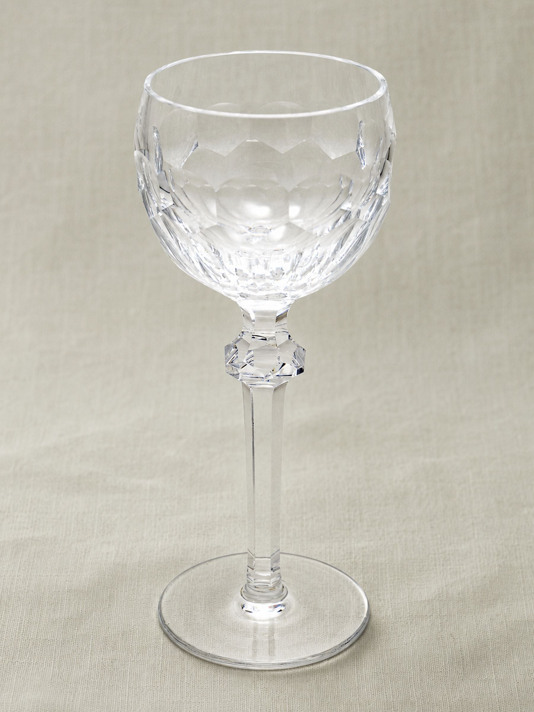 A clear cut-glass goblet with a faceted stem and circular base, photographed against a neutral beige fabric background.