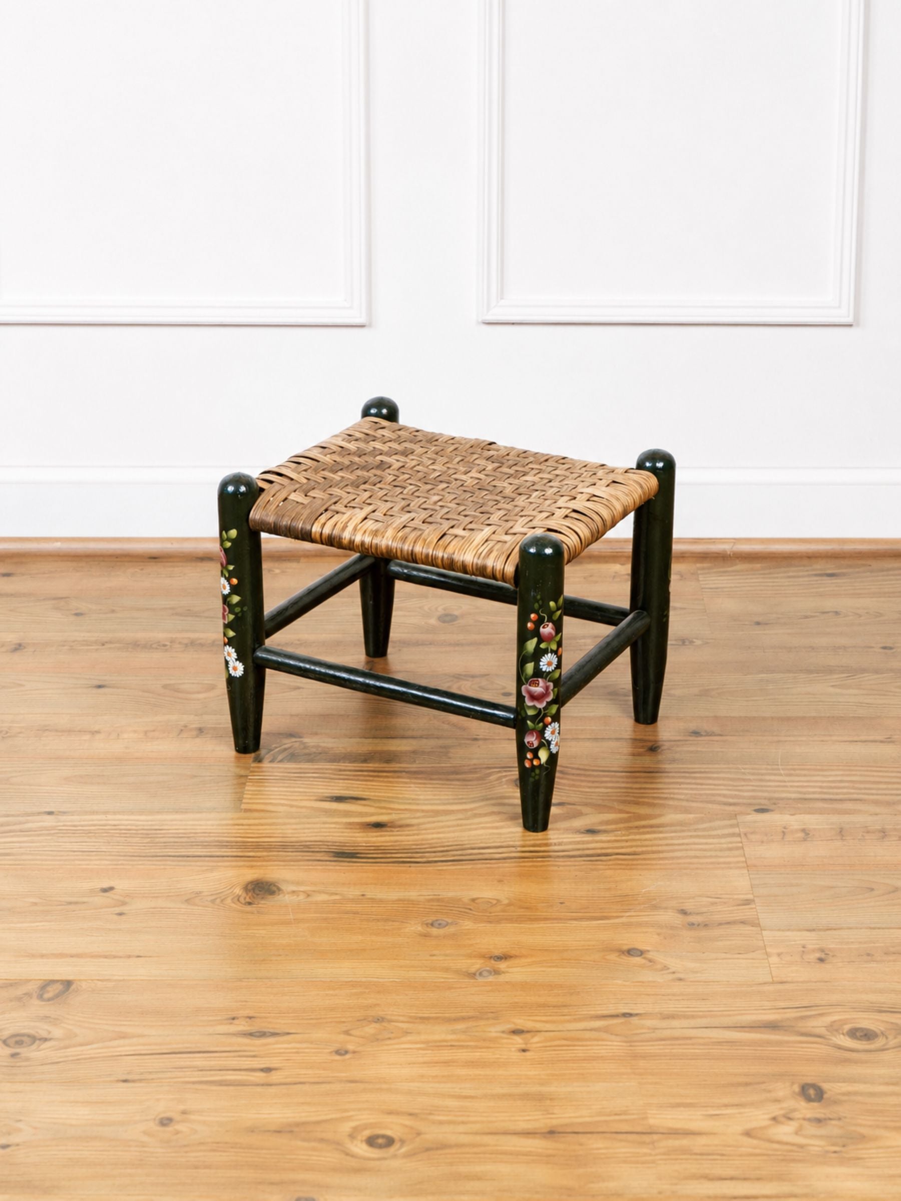 A small wooden stool with a woven straw seat and black painted legs decorated with colorful floral motifs, set on a light wood floor against a white paneled wall.