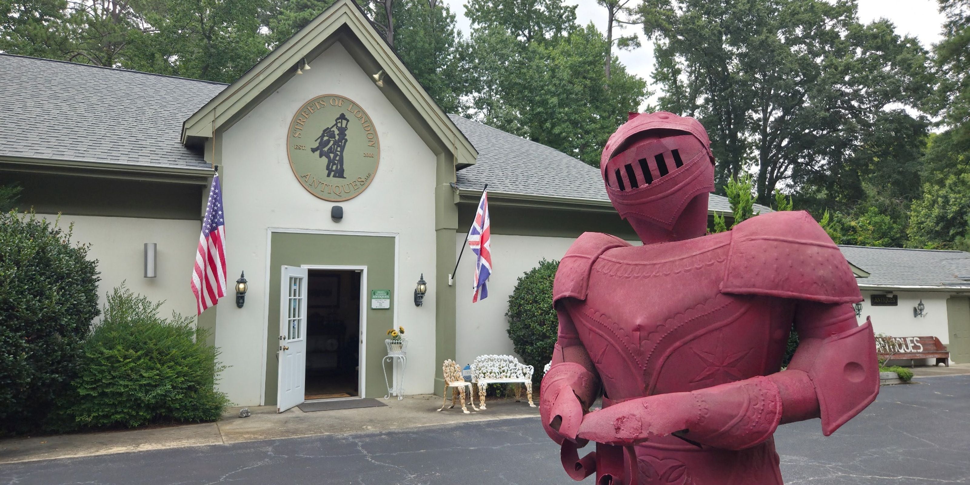 A large red knight statue in armor stands in front of a small building with a gray roof, trees, flags, and a driveway. The building has a circular emblem above the entrance and a sign on the right side.