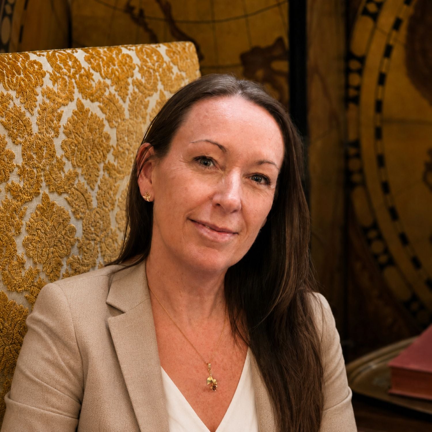A woman with long brown hair wearing a beige blazer and white top, seated in front of a gold and cream patterned armchair with a warm-toned decorative background.
