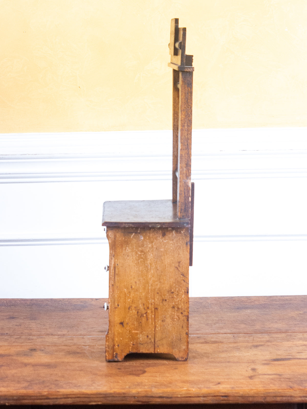 A vintage wooden chest of drawers with a tall, narrow back panel and a small metal drawer pull, shown standing on a wooden table against a yellow and white paneled wall.