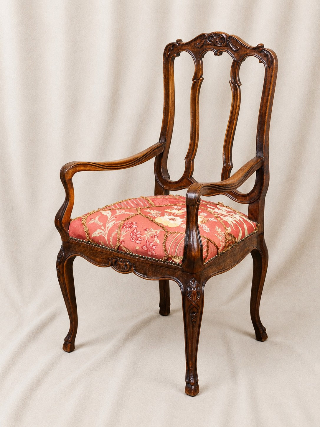 An antique-style wooden armchair with carved details, featuring a reddish-pink floral patterned upholstered seat cushion, photographed against a draped beige fabric backdrop.