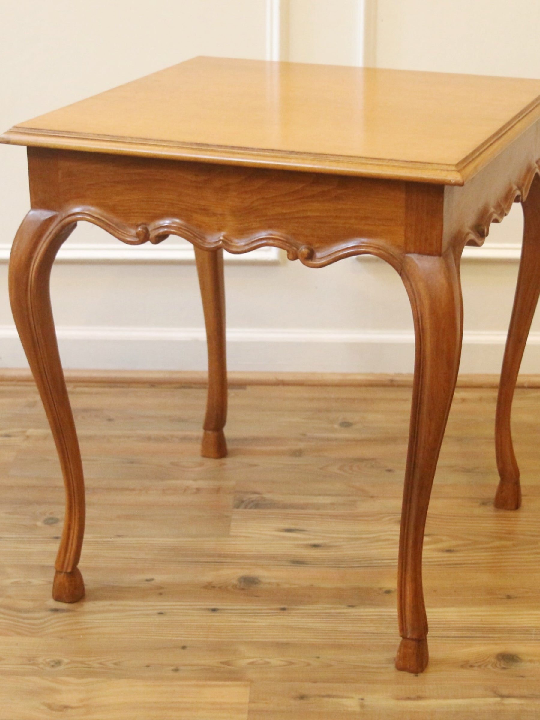 A square wooden end table with a warm honey finish and elegant cabriole legs, shown on a light wood floor against a white wall.