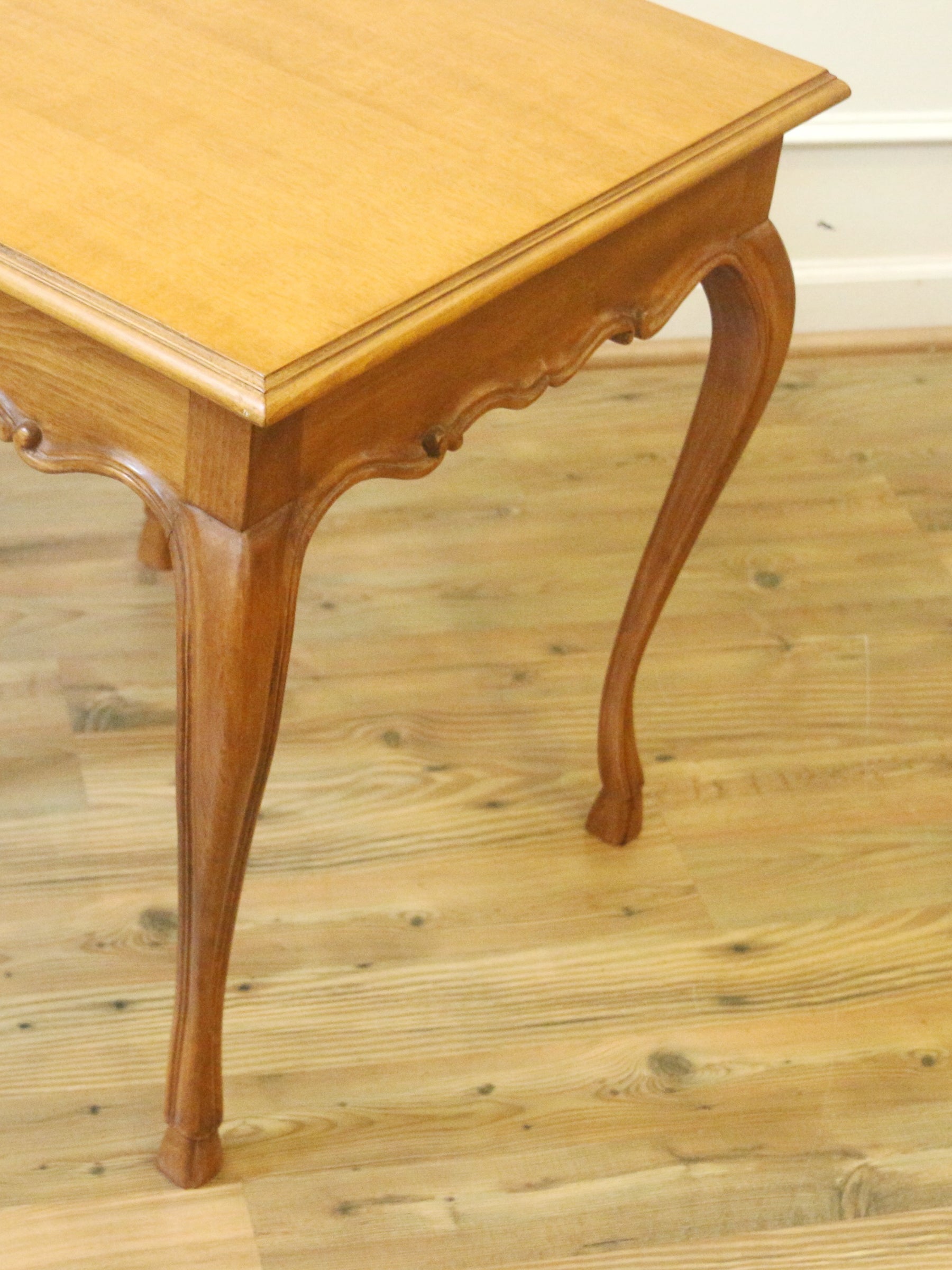 A close-up of a wooden table leg and corner, showing a warm honey-toned wood finish and curved cabriole-style legs on a light wood floor.
