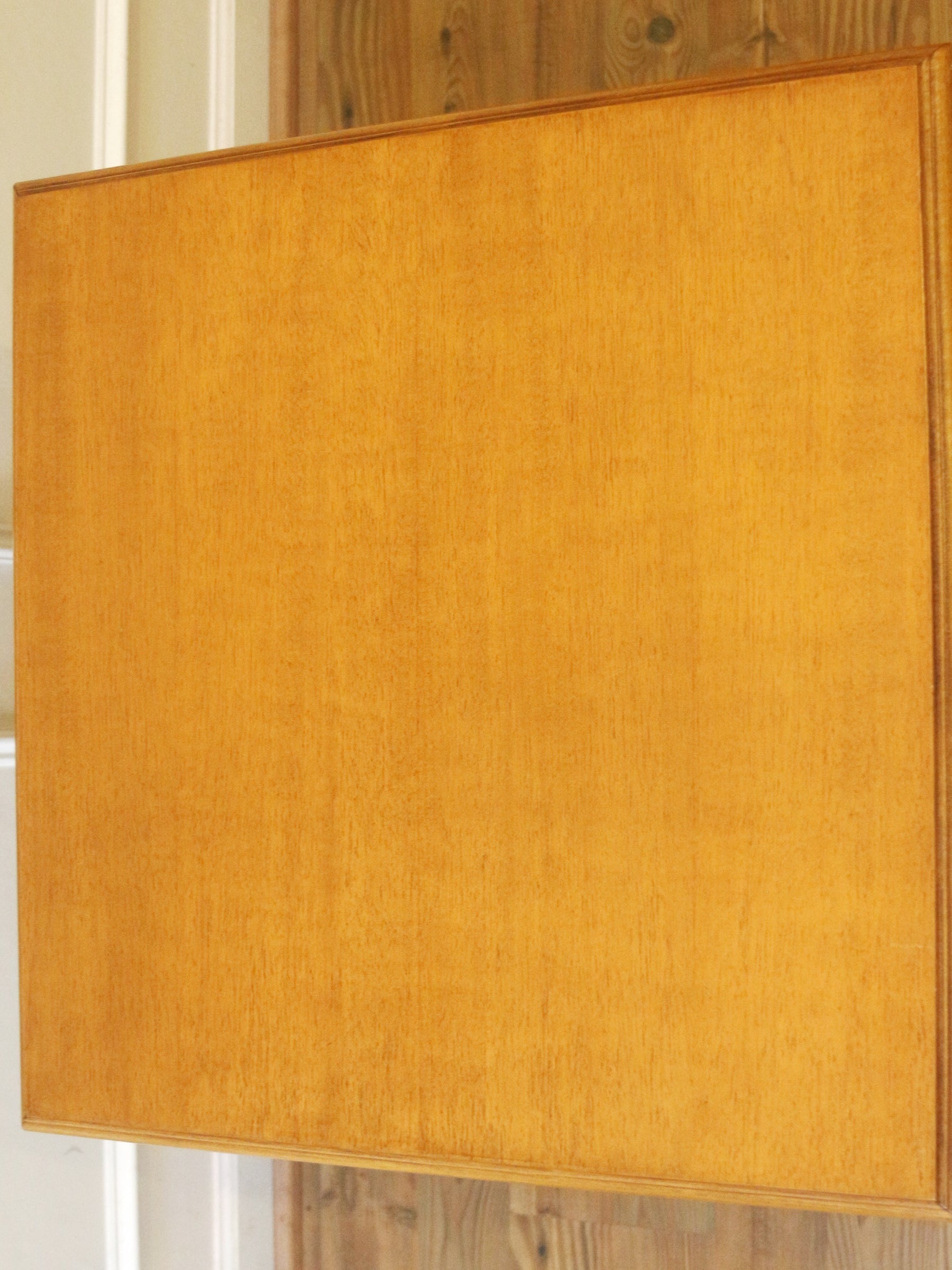 A square wooden panel or board with a warm honey-brown wood grain finish, shown against a background of white trim and wood flooring.