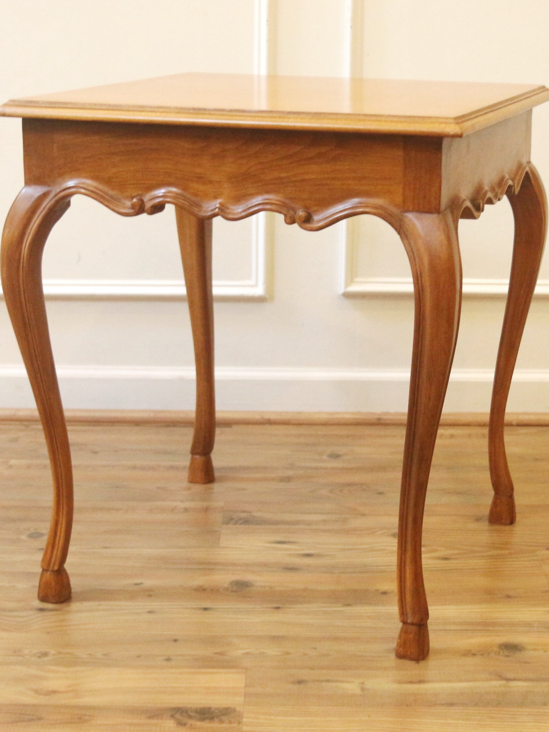 A wooden side table with a square top, carved cabriole-style legs, and a scalloped apron, shown on a light wood floor against a white paneled wall.