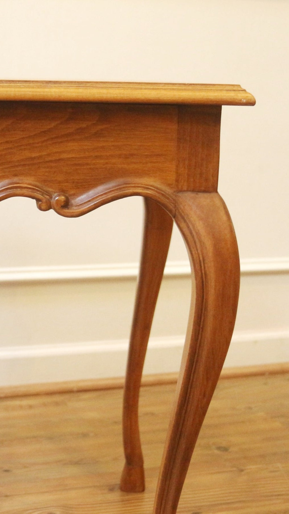 A wooden console table with curved cabriole-style legs and a decorative apron, shown in a warm medium brown finish against an off-white paneled wall and wood floor.
