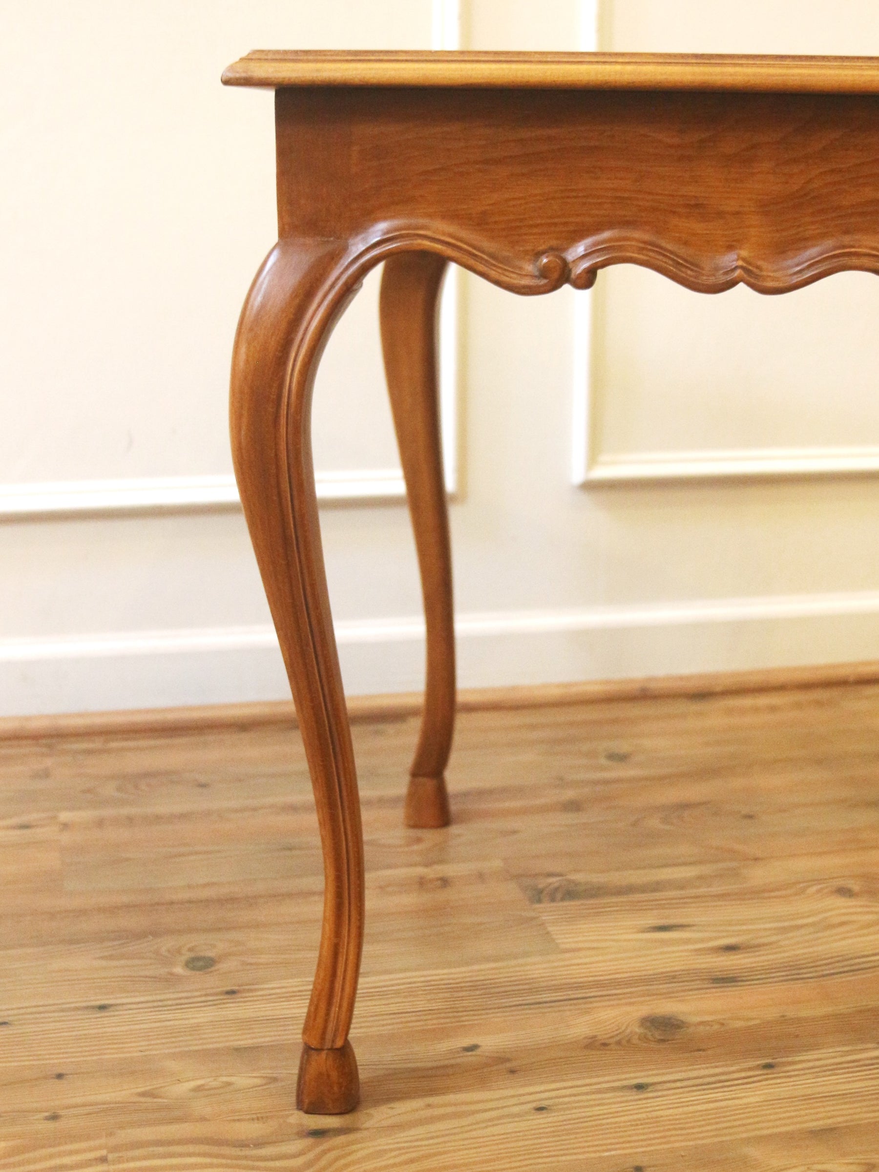 A close-up of a wooden console table leg with a curved cabriole design, set against a light cream wall and a light wood floor.