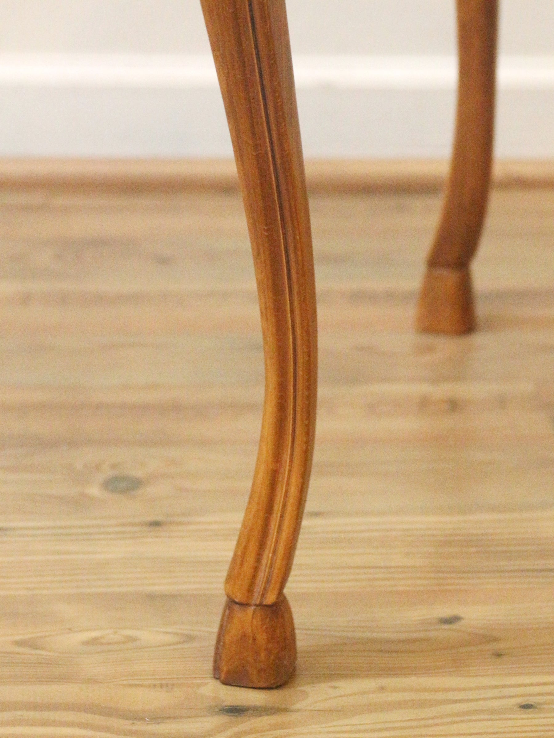 Close-up of a curved wooden table leg with a hoof foot, standing on a light wood floor.