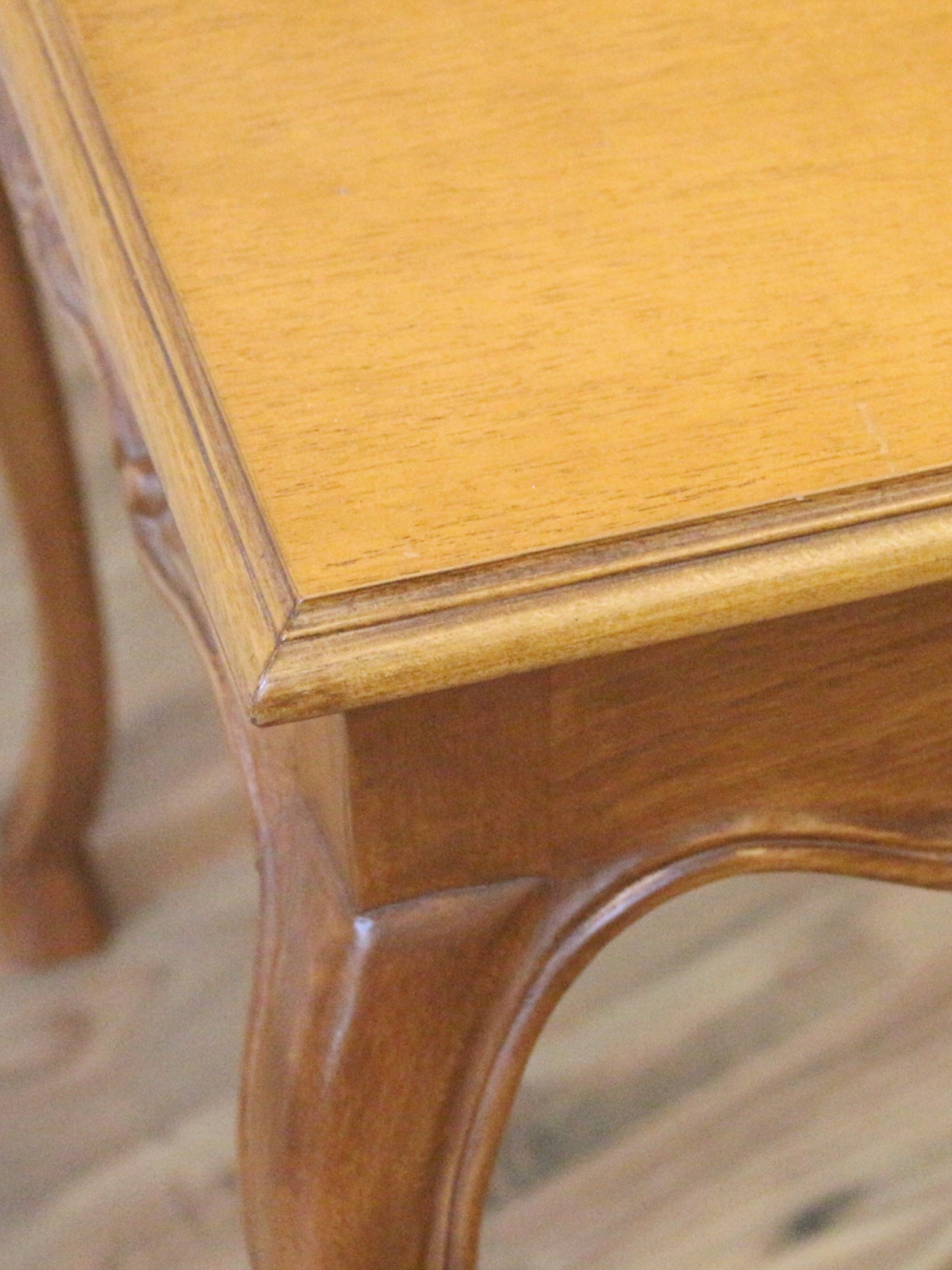 Close-up of a wooden table corner showing a light honey-toned top with a matching wood frame and curved legs on a light wood floor.