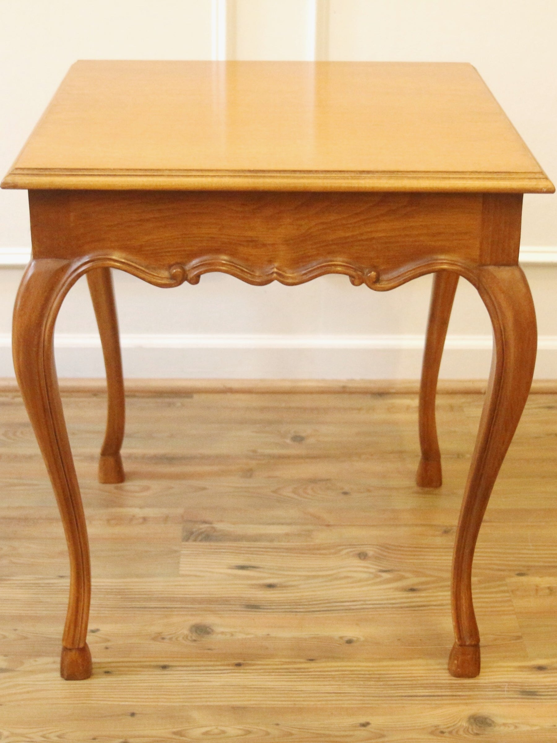 A square wooden end table with curved cabriole-style legs and a carved apron, shown on a light wood floor against a white wall.