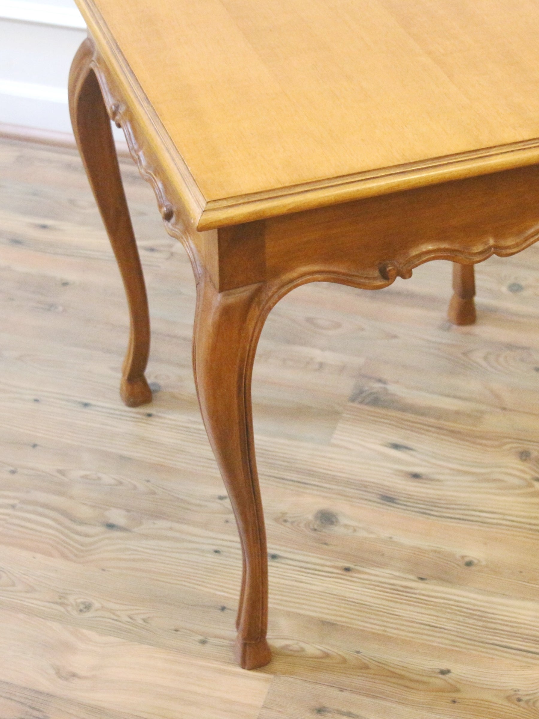 A close-up of a wooden dining table with curved cabriole-style legs and a light natural wood top, shown on a light wood plank floor.