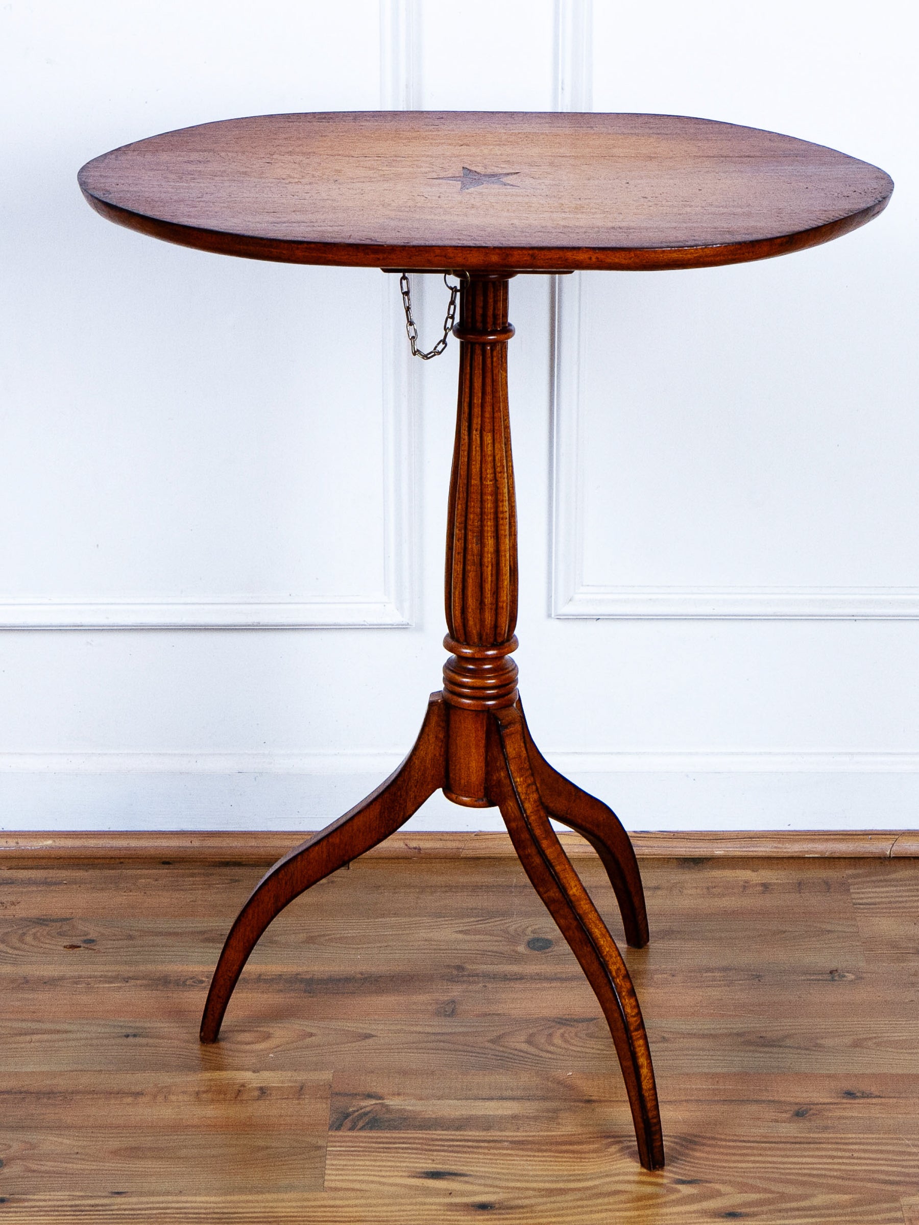 A round wooden pedestal tilt-top table shown with the top horizontal with a three-legged base, shown against a white paneled wall and wood floor.