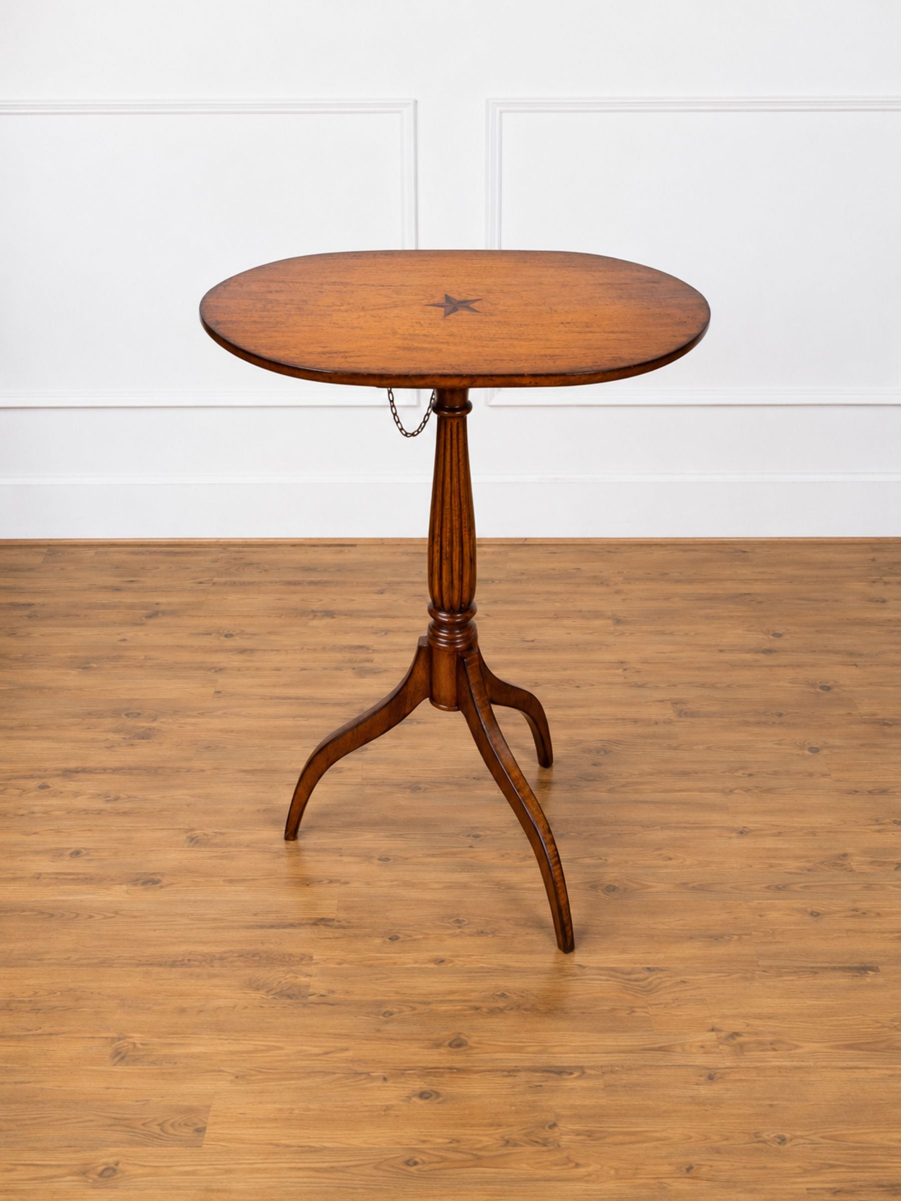 A small round wooden table with a turned pedestal base and three curved legs, shown on a wood floor against a white paneled wall.