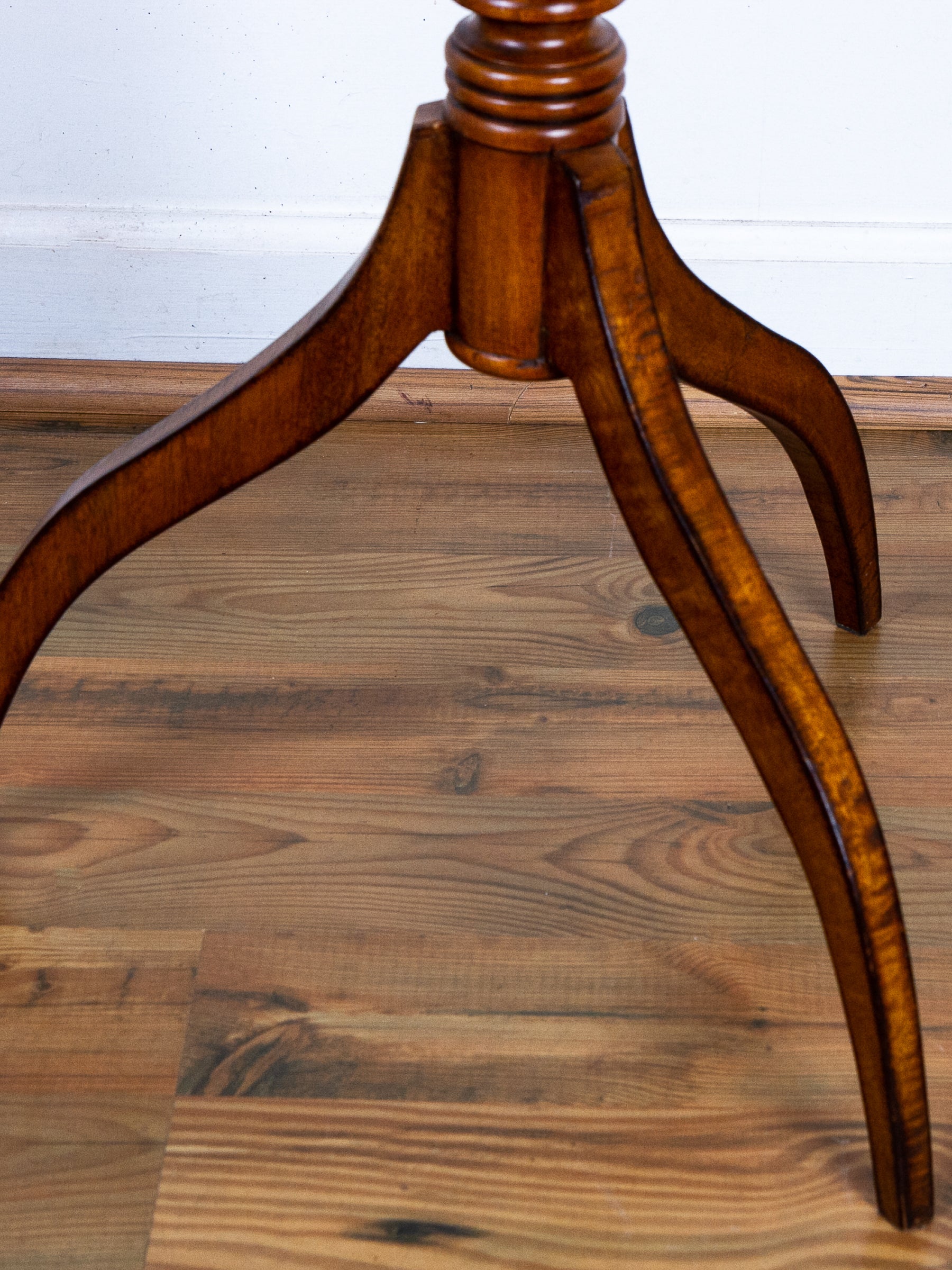 Close-up of a polished wooden table base with three curved legs on a wood floor against a white wall.