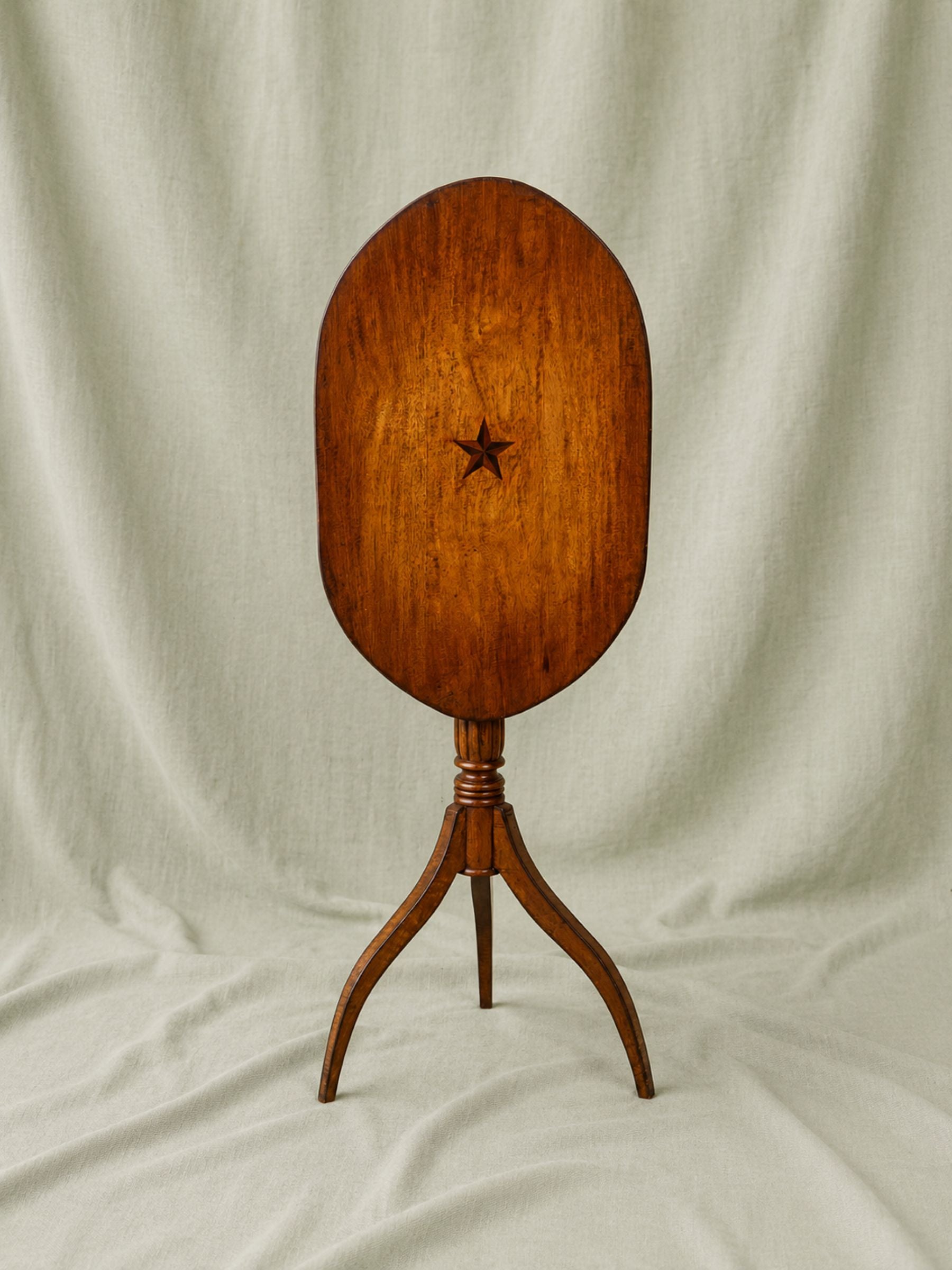 A vintage wooden table with an oval tilt-top tabletop featuring a star inlay, supported by a three-legged pedestal base, set against a draped cream fabric backdrop.