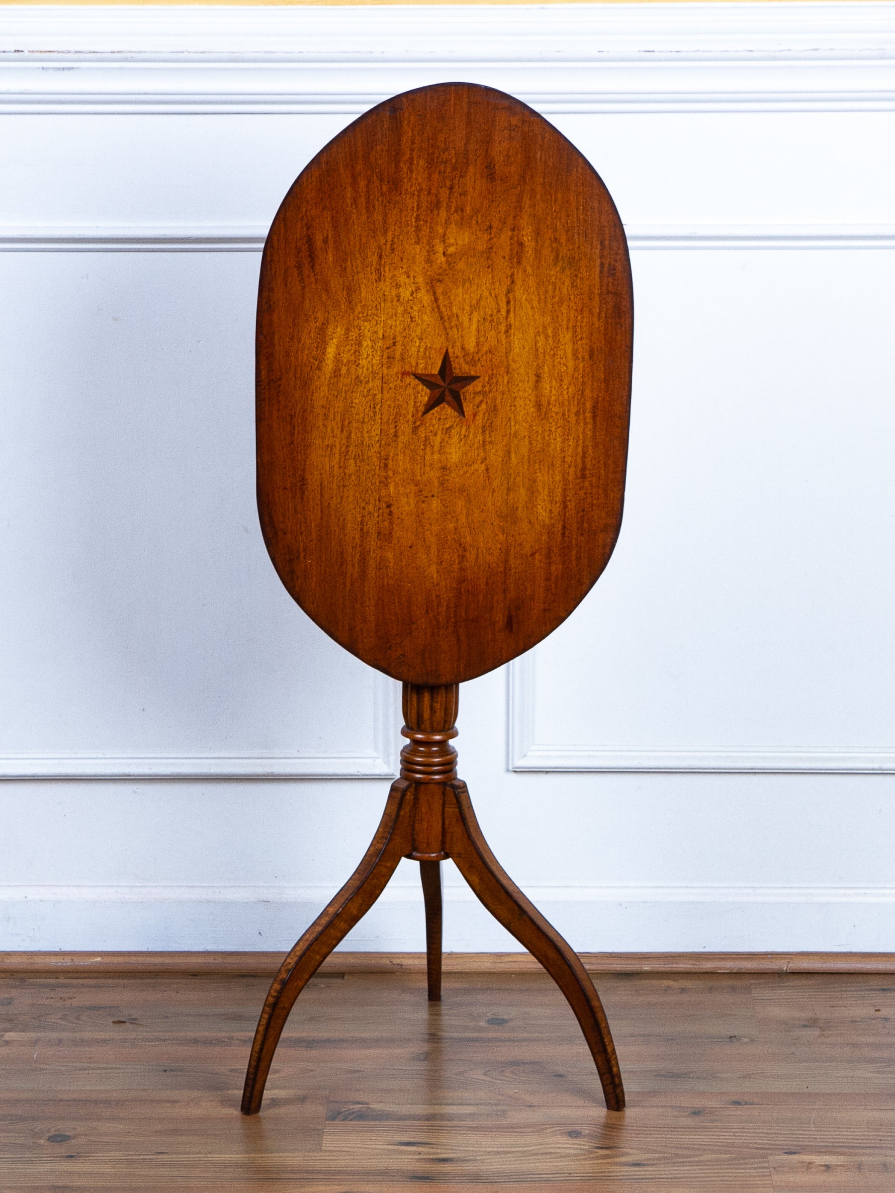An antique wooden oval tilt-top table with a star inlay on the front of the leaf, supported by a three-legged pedestal base, set against a white paneled wall and wooden floor.