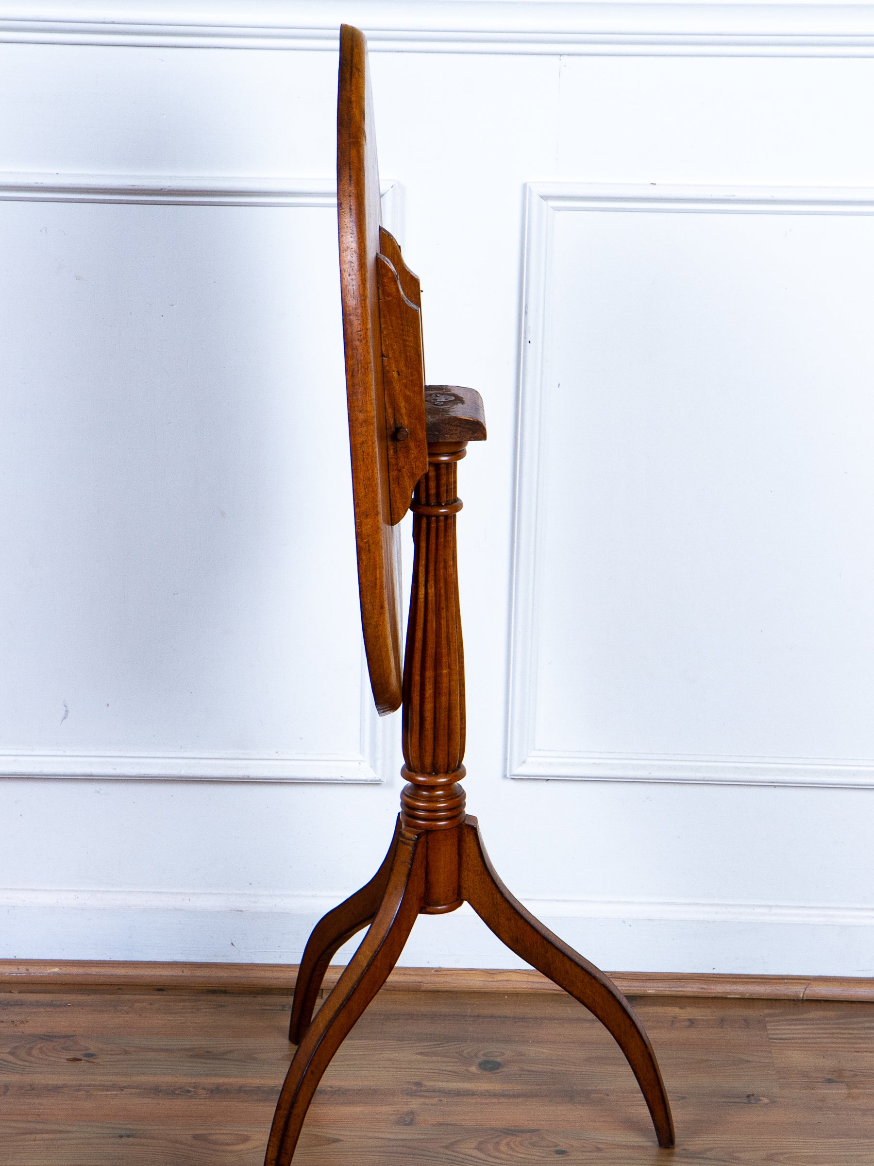 A vintage wooden tilt-top table with a round top, turned legs, and a central pedestal base, shown from a side profile against a white paneled wall and wood floor.