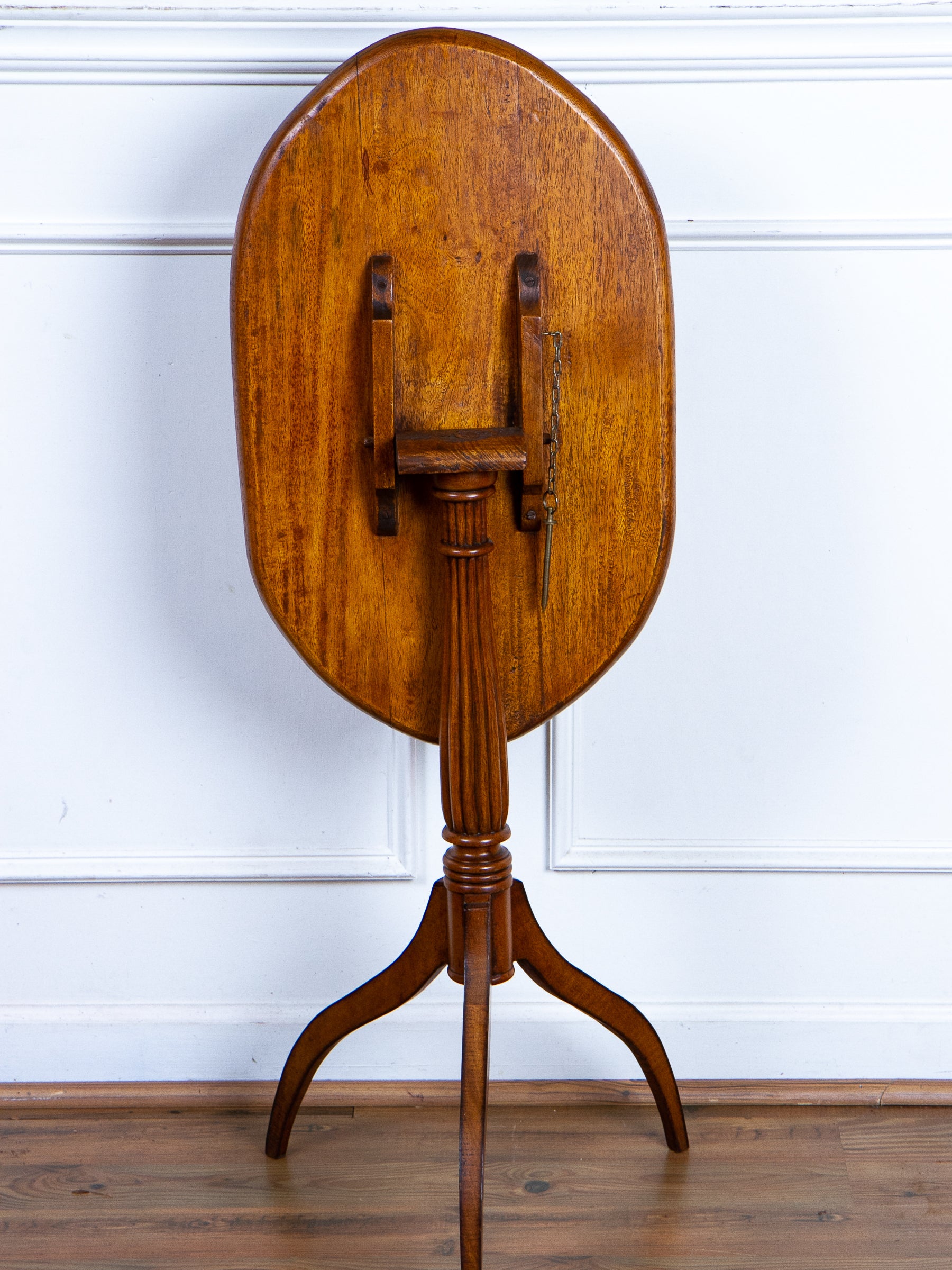 An antique wooden oval tilt-top table shown from the back on a three-legged pedestal stand, photographed against a white paneled wall and wood floor.
