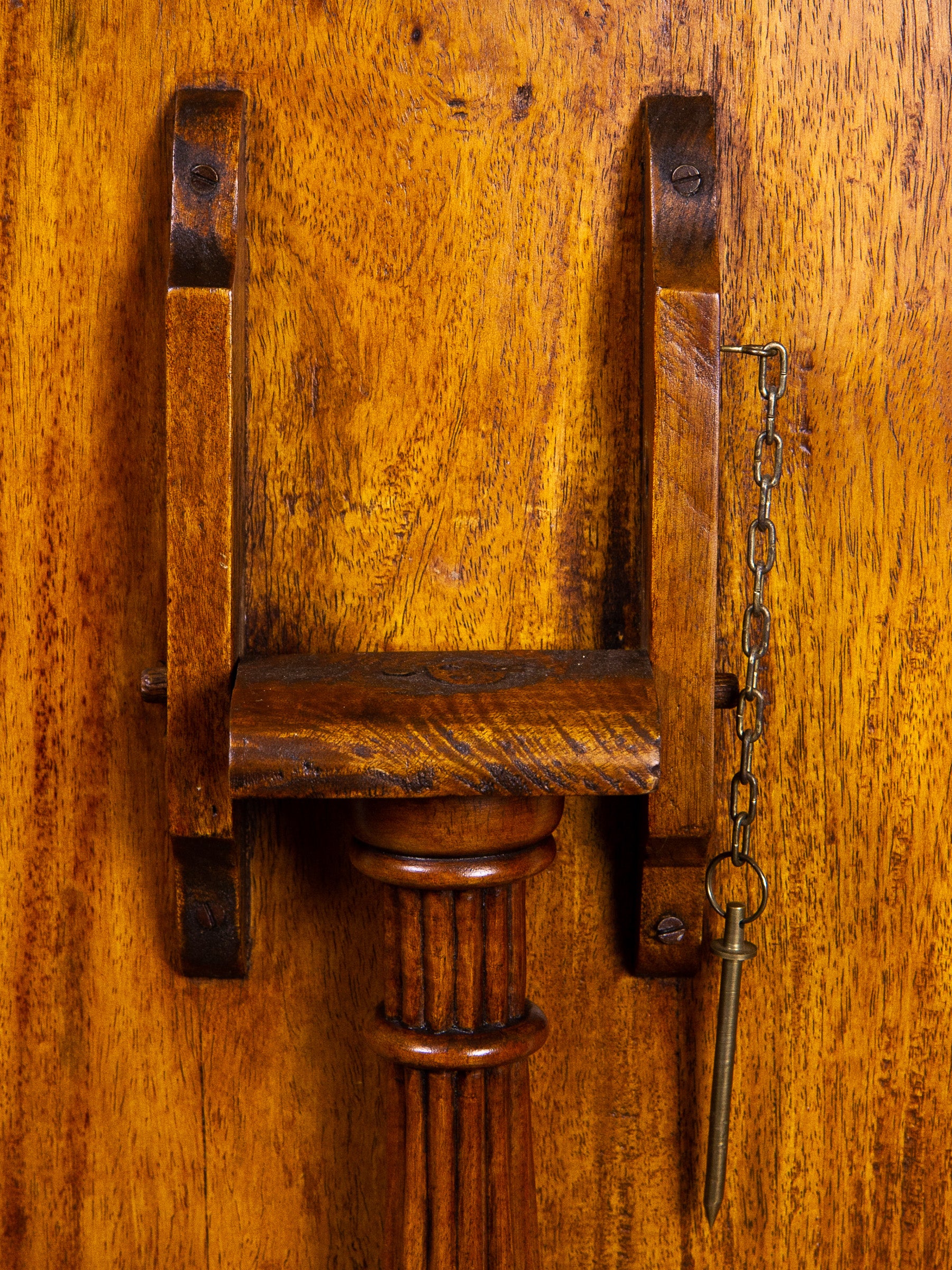 Close-up of a vintage wooden tilt-top table lock mechanism with the fluted column top of the pedestal, crosspiece, and hanging chain with key on a warm wood panel background.