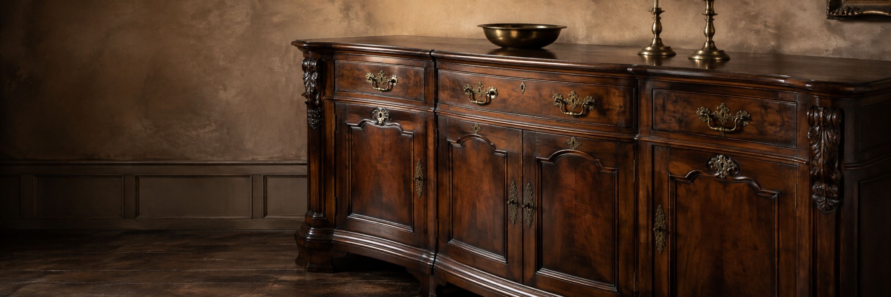 Antique wooden sideboard with paneled doors, ornate brass hardware, candlesticks and a decorative brass bowl.