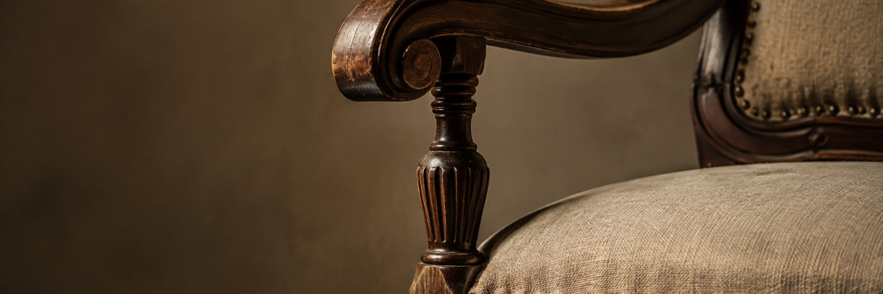 Close-up of an antique carved wood armchair with neutral linen upholstery and brass nailhead trim.