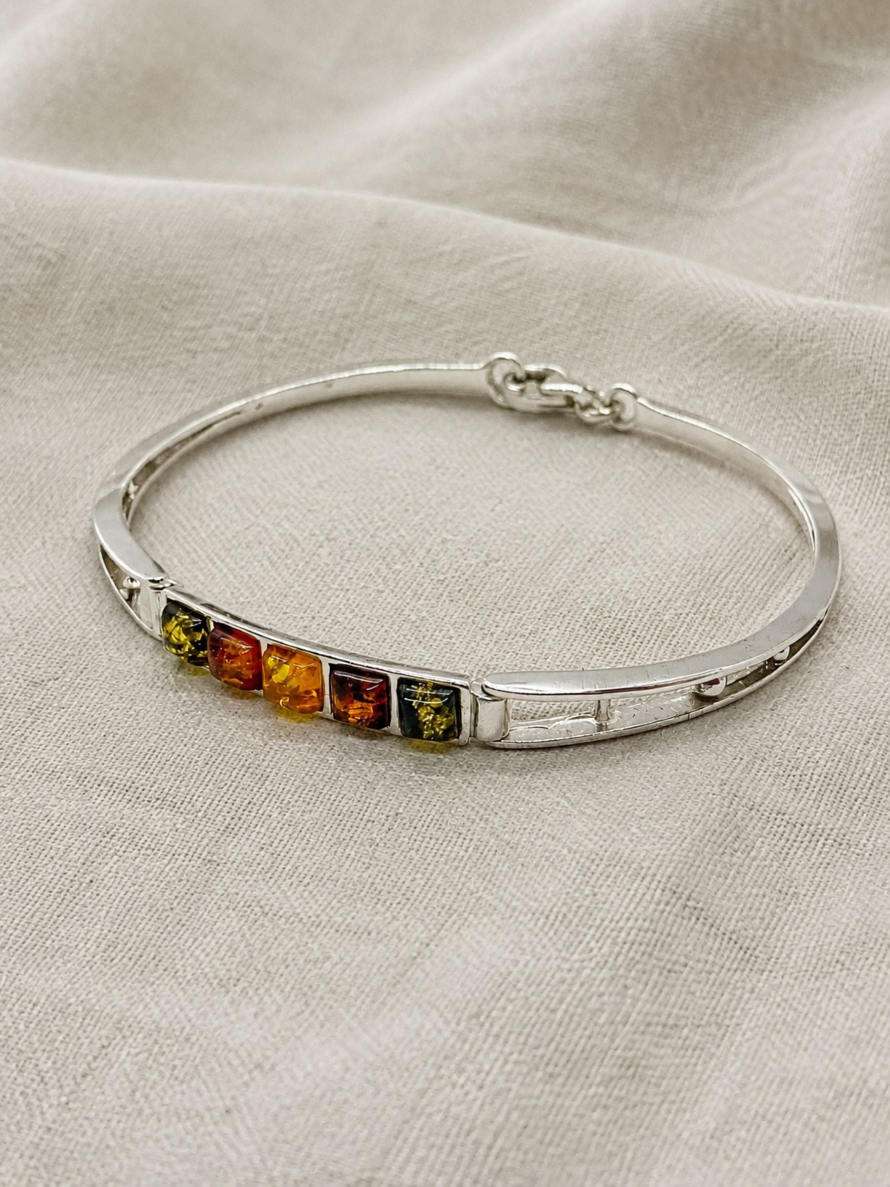 A polished sterling silver bangle bracelet featuring five small square-cut amber stones in yellow, orange, and green hues, displayed on a textured beige linen fabric background.