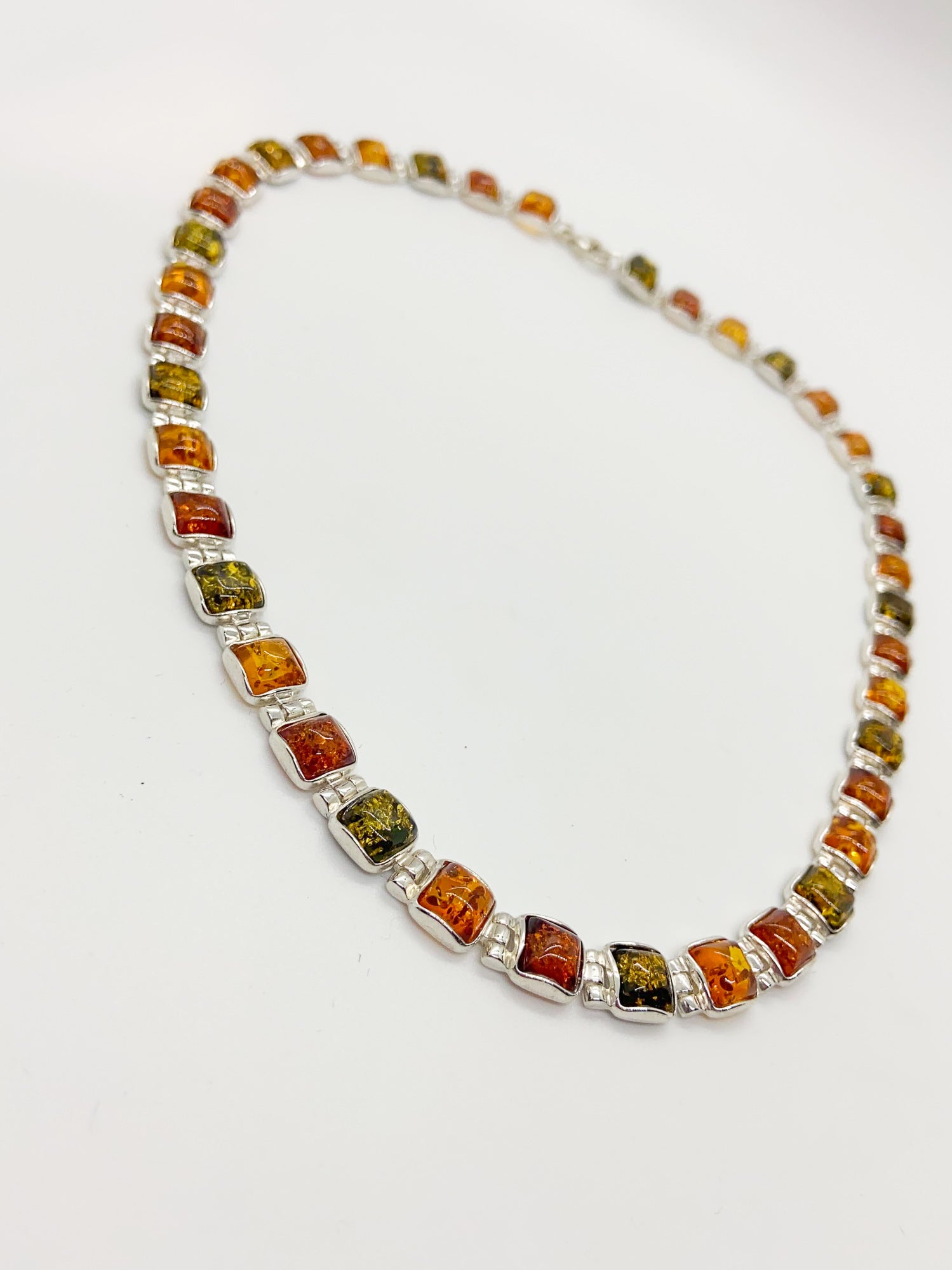 A sterling silver beaded necklace featuring square genuine amber stones in warm orange, brown, and olive green hues, arranged in a circular shape on a plain white background.