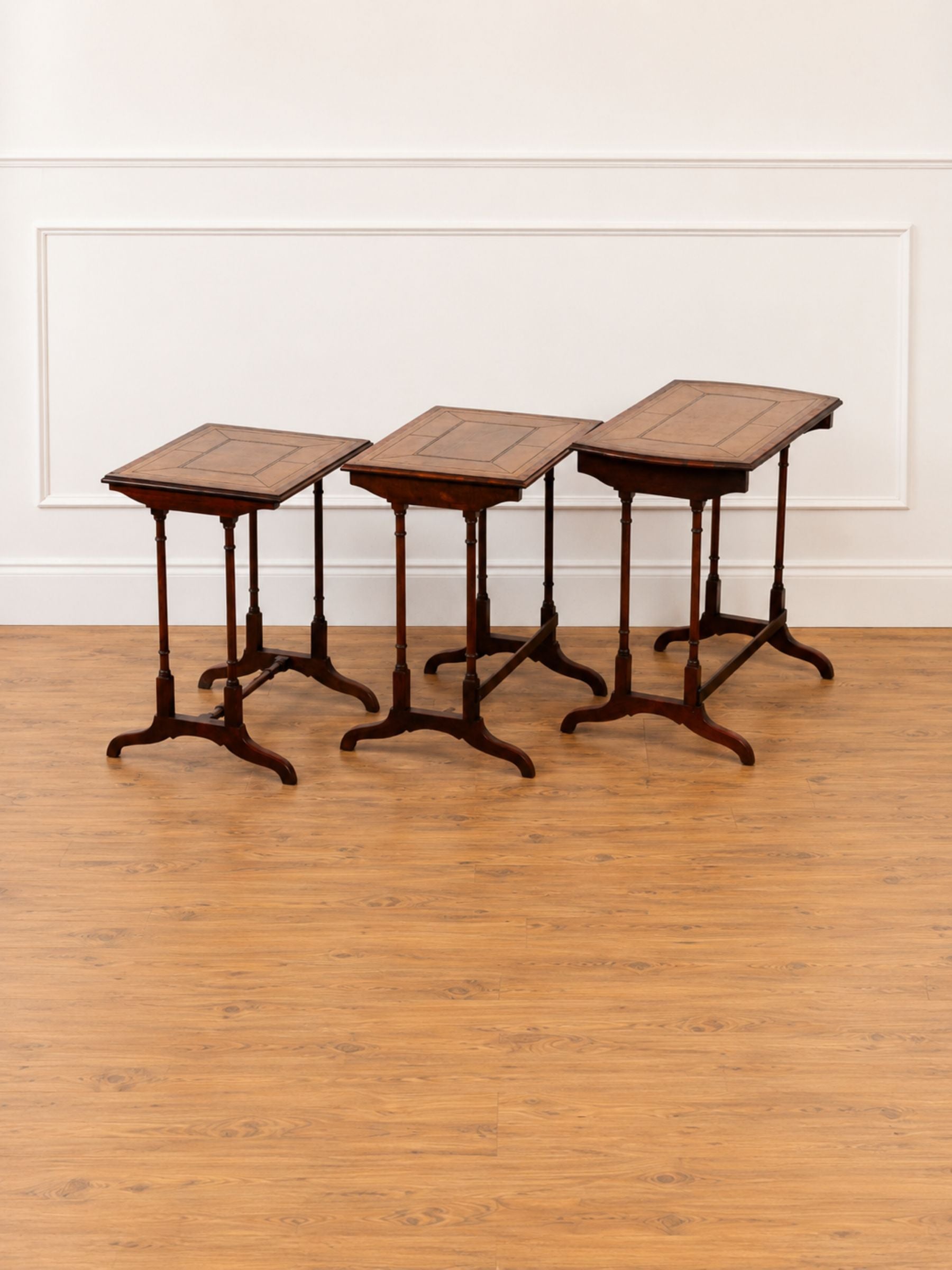 Three nested wooden nesting tables with dark brown wood grain and olive green leather tops, set on a light wood floor against a white paneled wall.