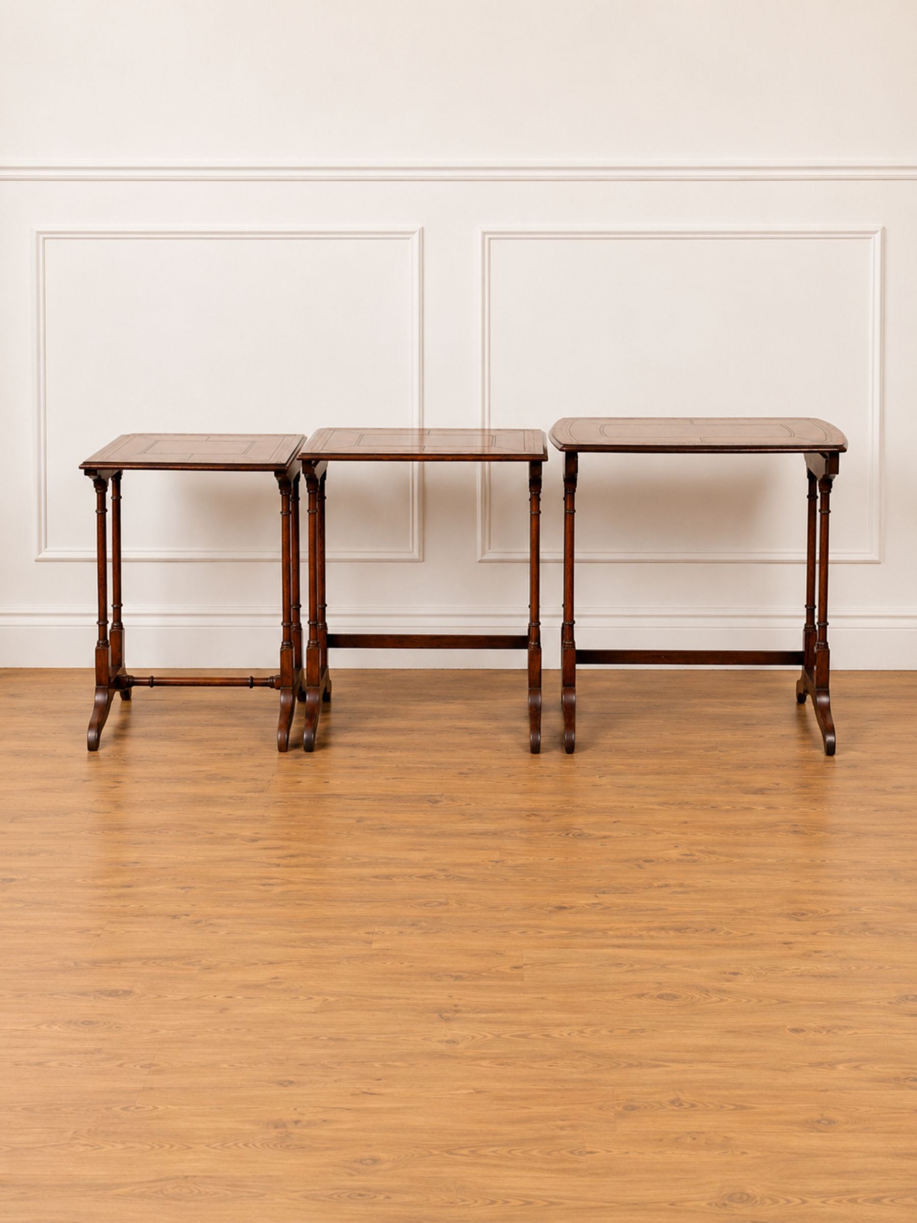 A set of three matching wooden nesting tables or side tables with square brown tops and dark brown turned legs, arranged against a white paneled wall on a light wood floor.