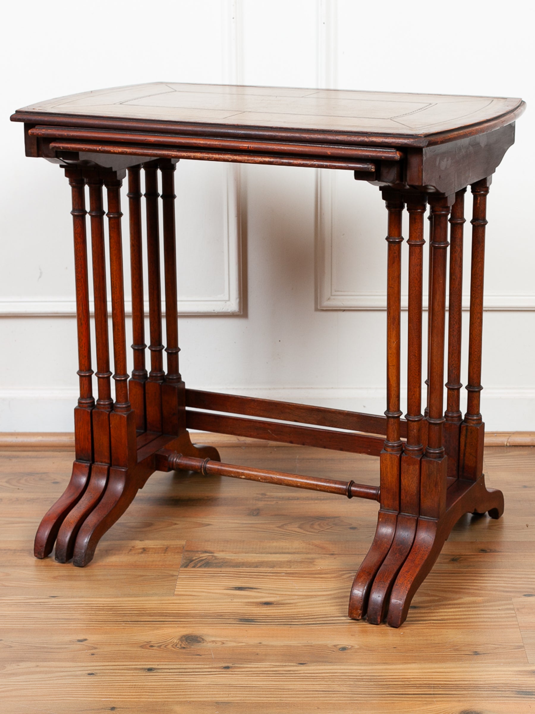 An antique dark mahogany wood set of 3 nesting tables with turned spindle legs and a central stretcher bar, shown on a wooden floor against a white paneled wall.