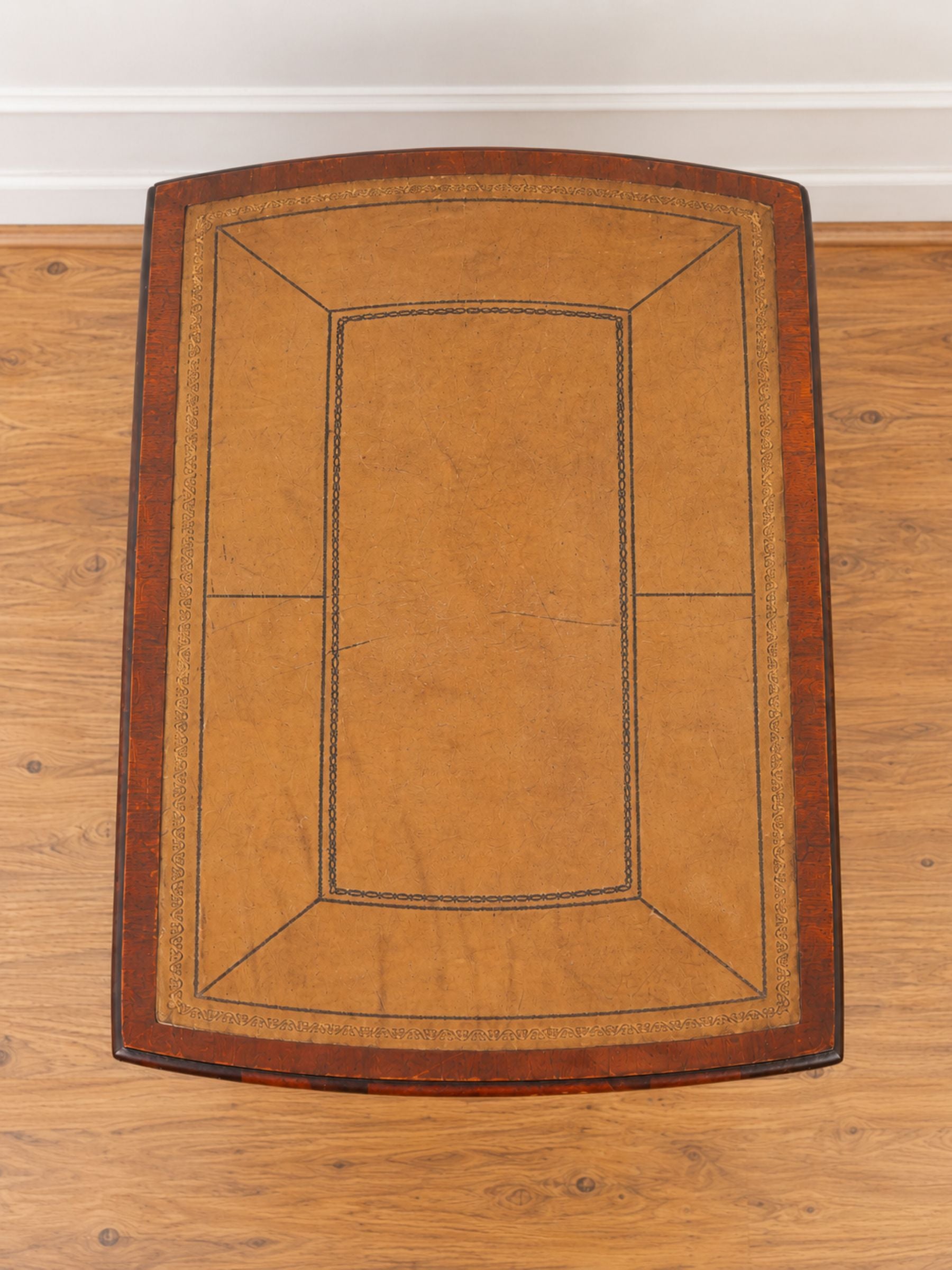 A rectangular wooden table top viewed from above, featuring a warm tan leather inlay with dark stitched geometric lines and a dark brown wood frame, set on a light wood floor.