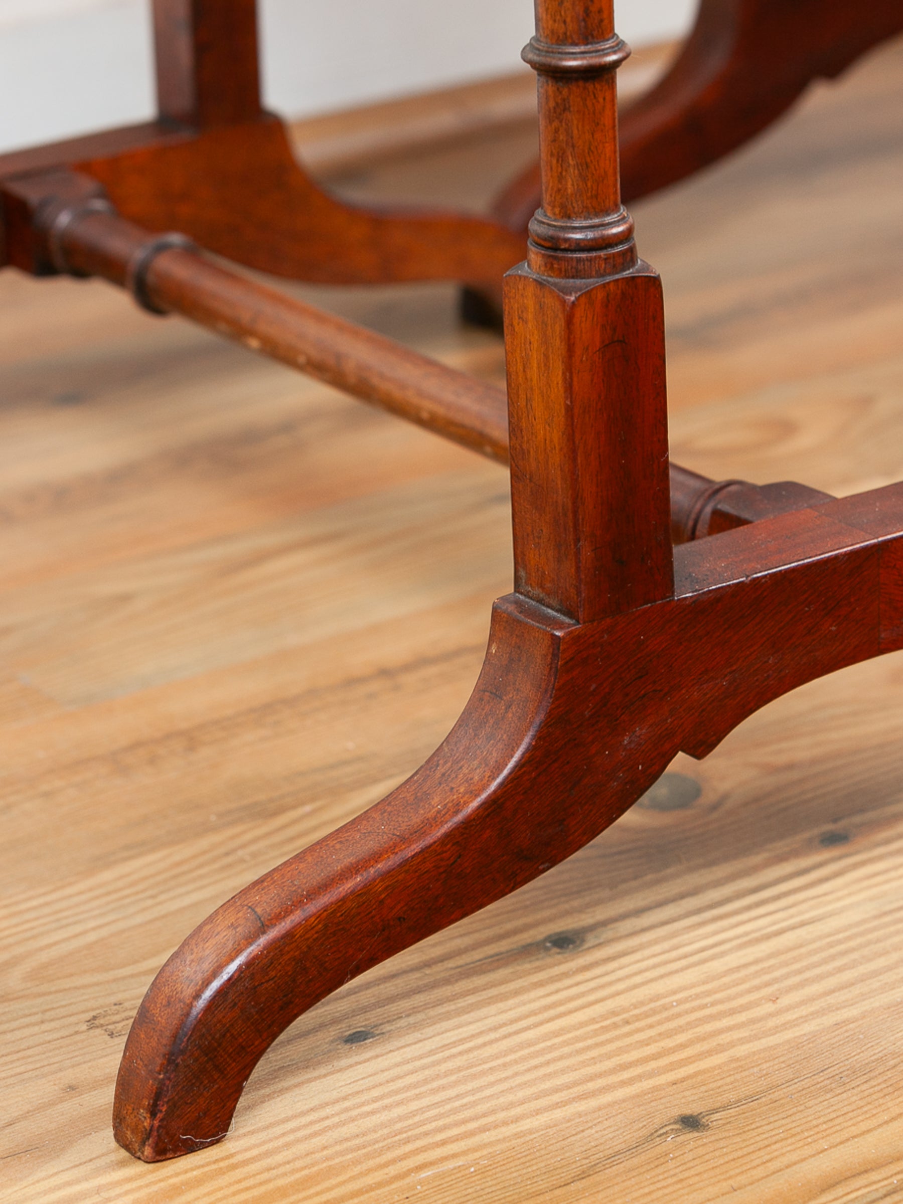 A close-up of a dark reddish-brown wooden table leg with a curved foot and central support post, resting on a light wood floor.
