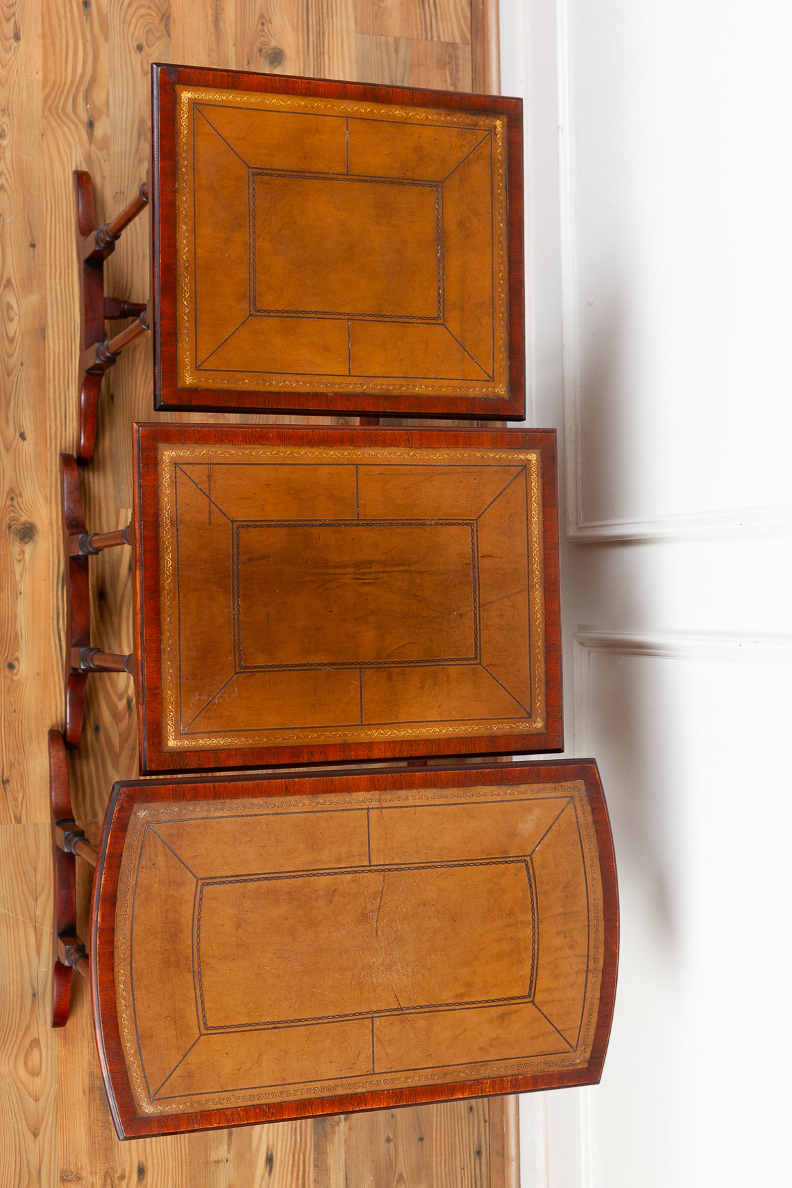 Three antique wooden nesting tables from above, showcasing warm olive toned leather tops with dark brown and gold geometric inlay detailing, set against a background of a wooden floor and white wall.